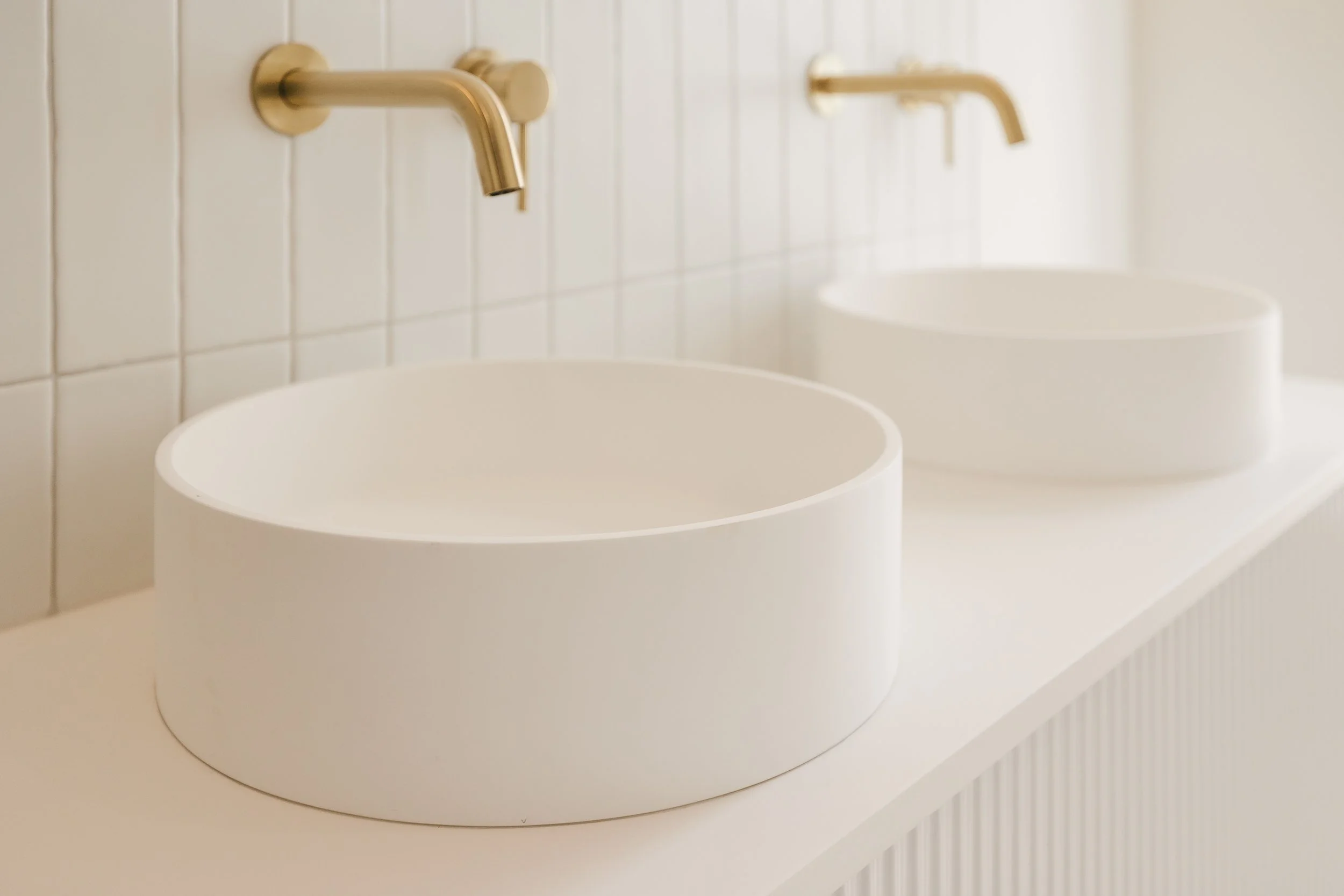 Two white ceramic sinks with gold wall-mounted faucets in a bathroom with white tiled wall.