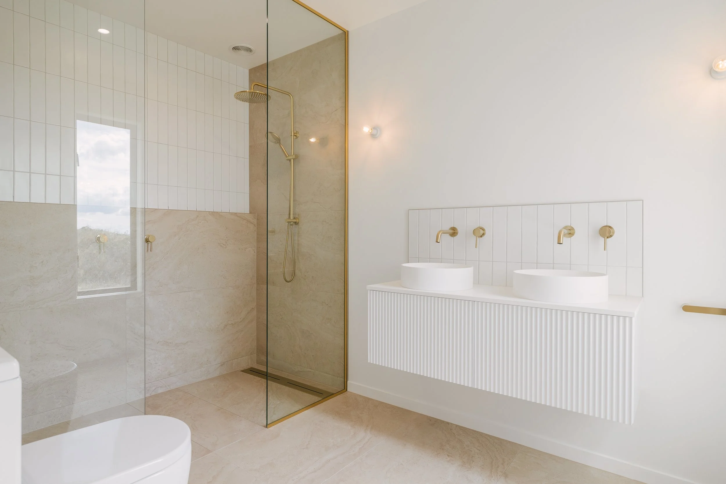 Modern bathroom featuring a walk-in shower with a glass door, gold fixtures, and beige tiles. There are two white vessel sinks with gold faucets on a white ribbed vanity, and wall-mounted lights on a white wall.