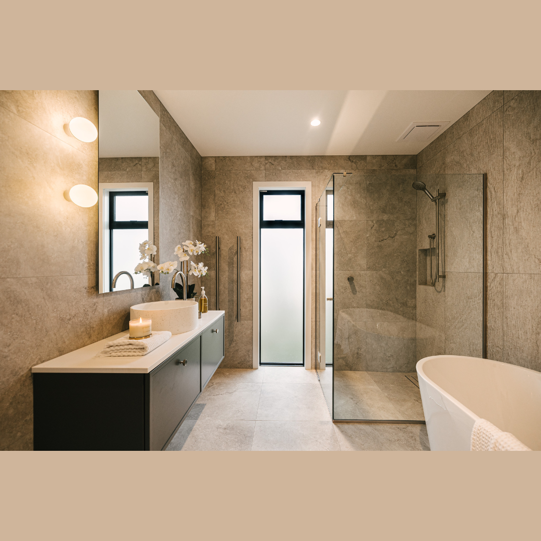 Modern bathroom with beige tiled walls and floor, a dark vanity with a white countertop, a circular vessel sink, a large mirror, two small frosted windows, a frosted glass door leading to a shower area, and a free-standing bathtub.