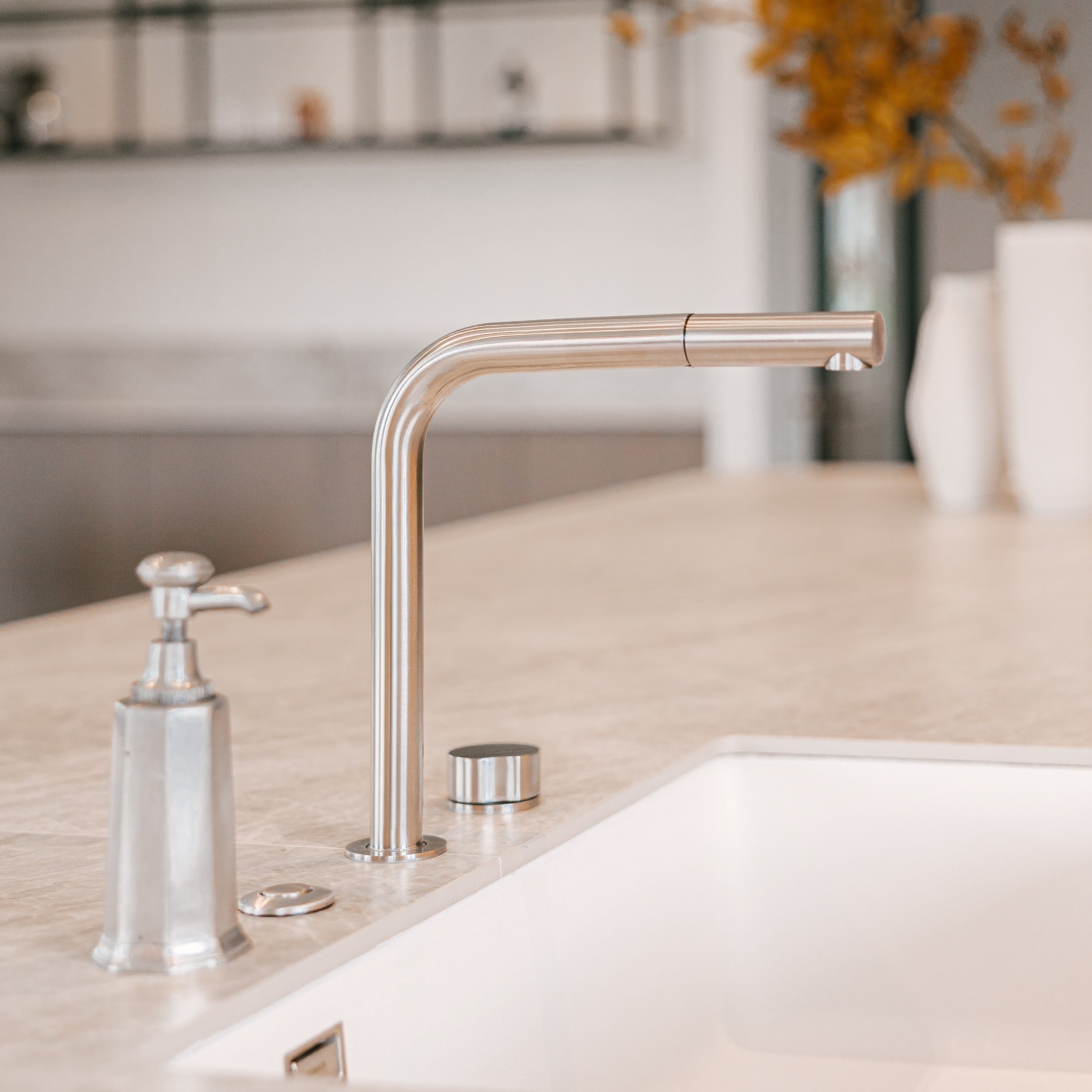 Close-up view of a modern kitchen faucet with a beige countertop and a blurred background featuring framed pictures, a white vase with orange flowers, and a window.