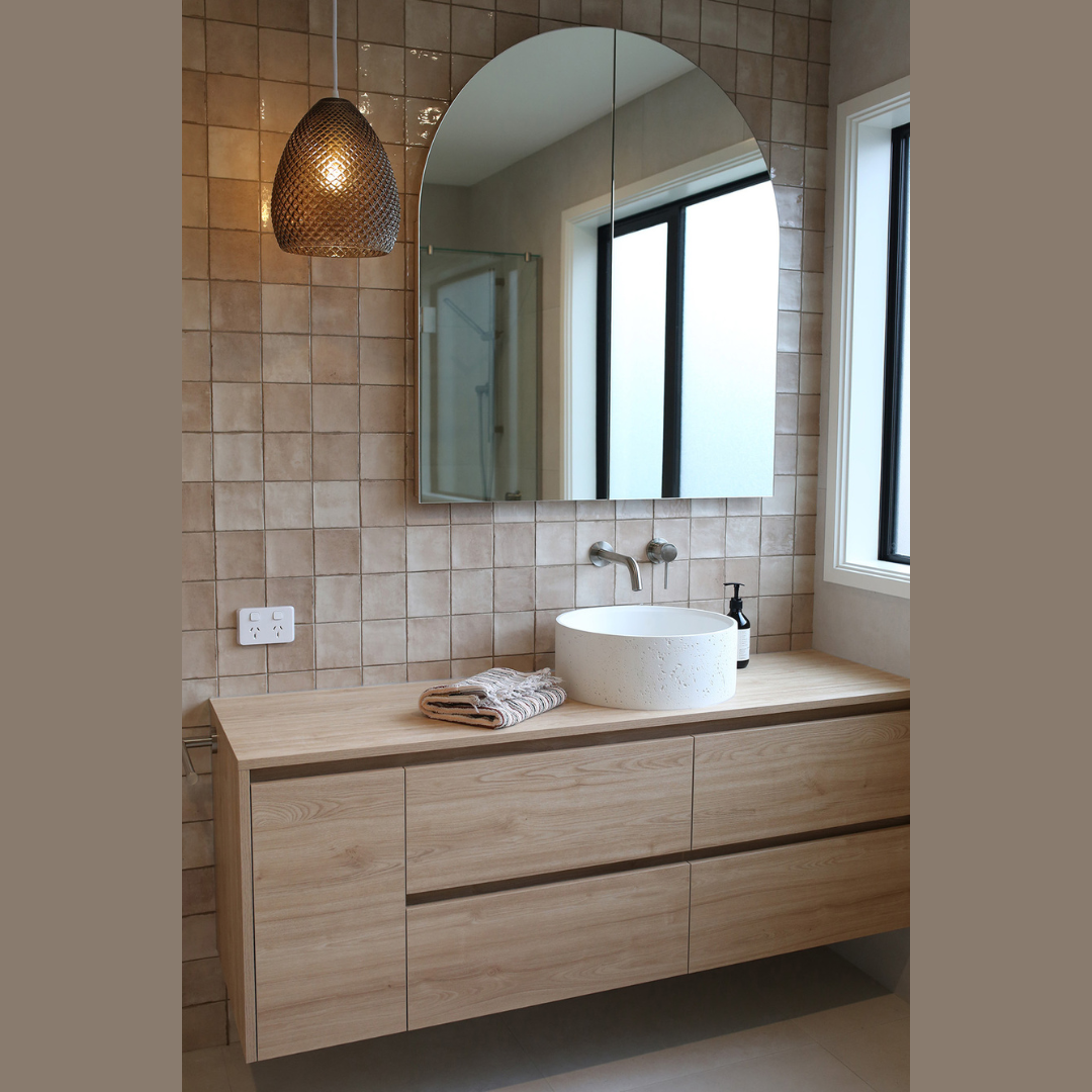 Modern bathroom with beige tiled wall, wooden vanity with three drawers, oval vessel sink, wall-mounted faucet, large mirror, window, and pendant light.