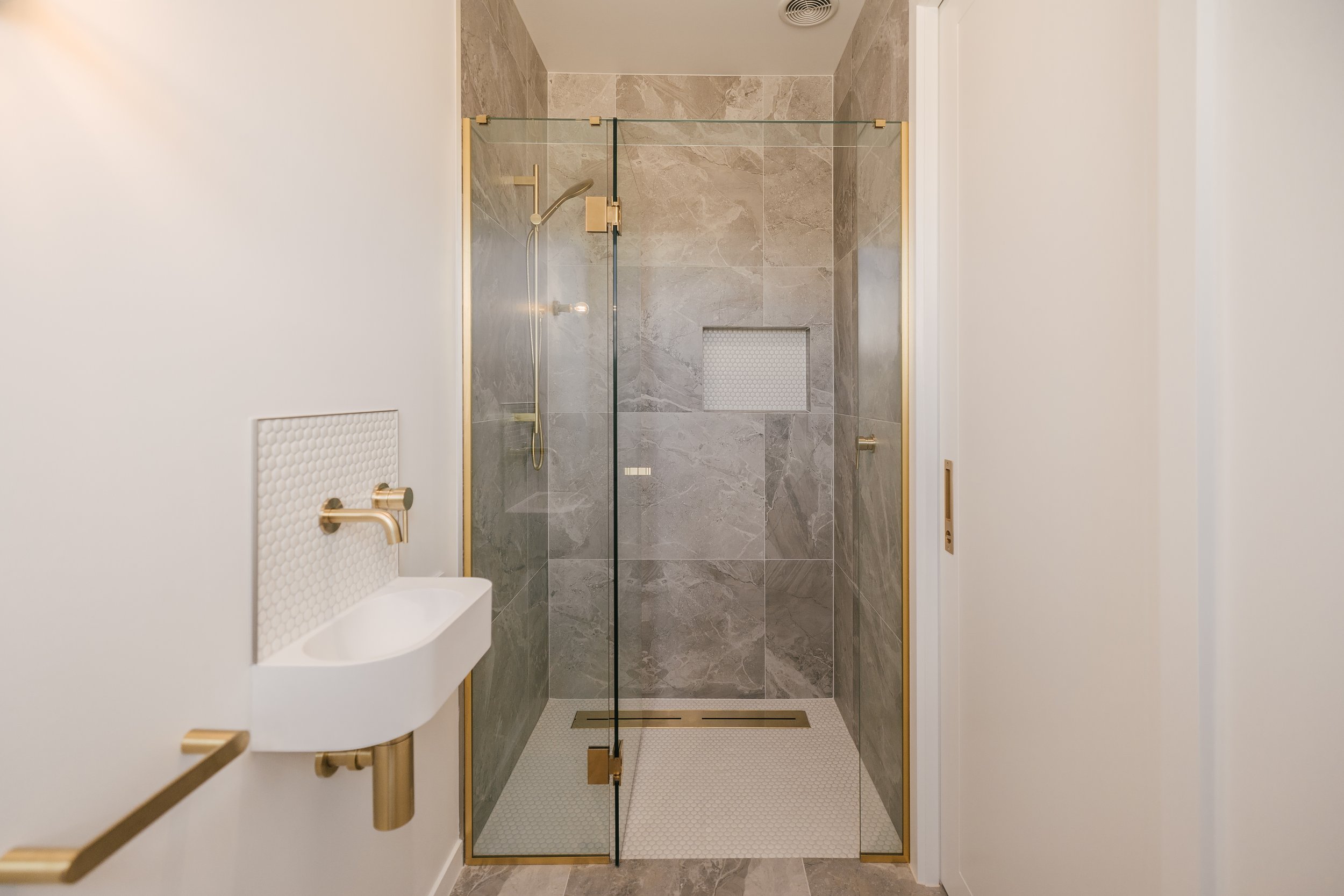 Modern walk-in shower with beige marble tiles, glass door with gold hardware, and a built-in niche. White walls with textured hexagonal tile accents near the sink, gold fixtures, and part of a white countertop are also visible.