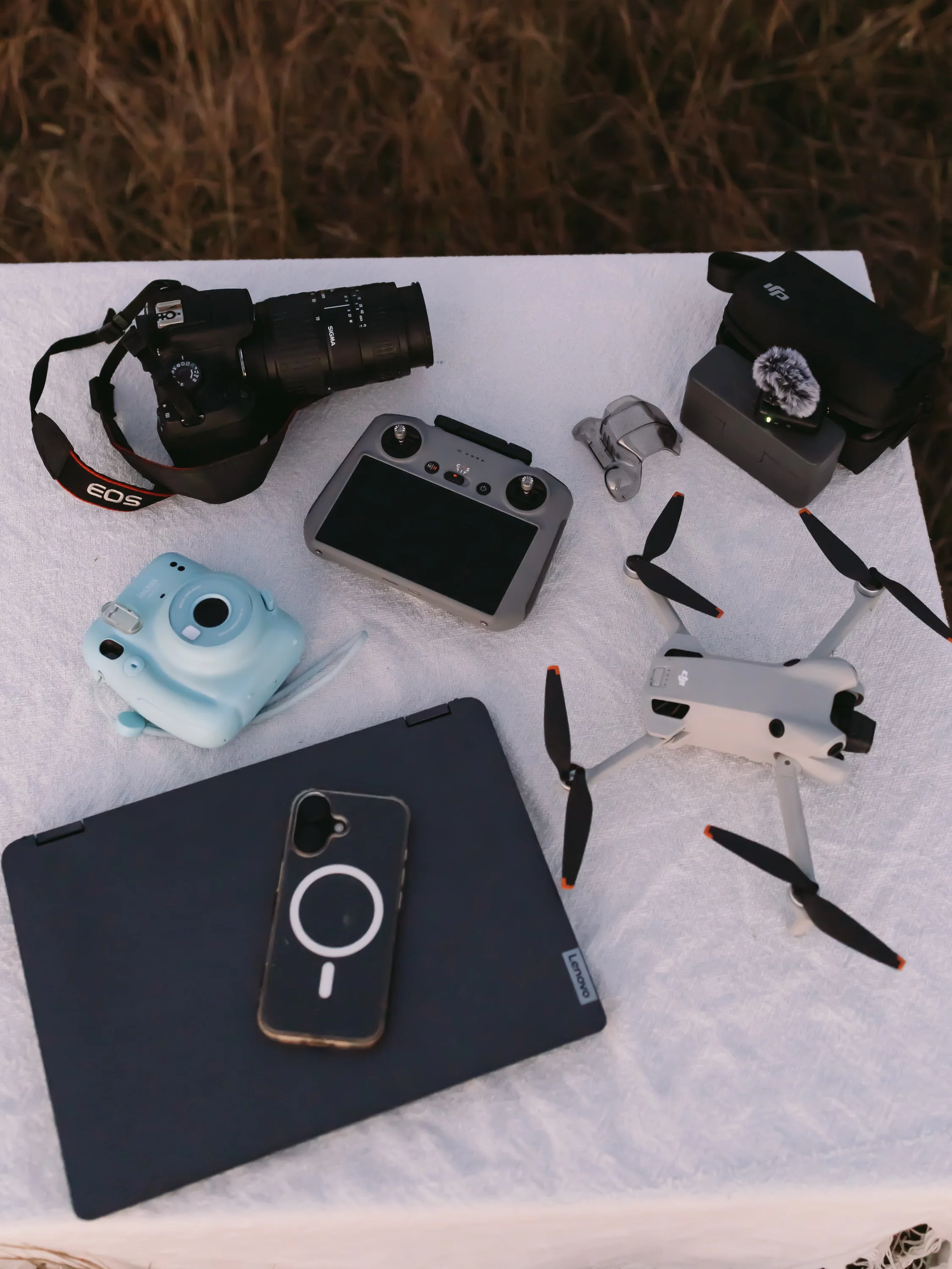 A collection of photography equipment on a white surface: a Canon EOS camera with a long zoom lens, a drone, a remote control, a small camera, a black tablet, a camera case with a furry microphone, and a protective camera cover.