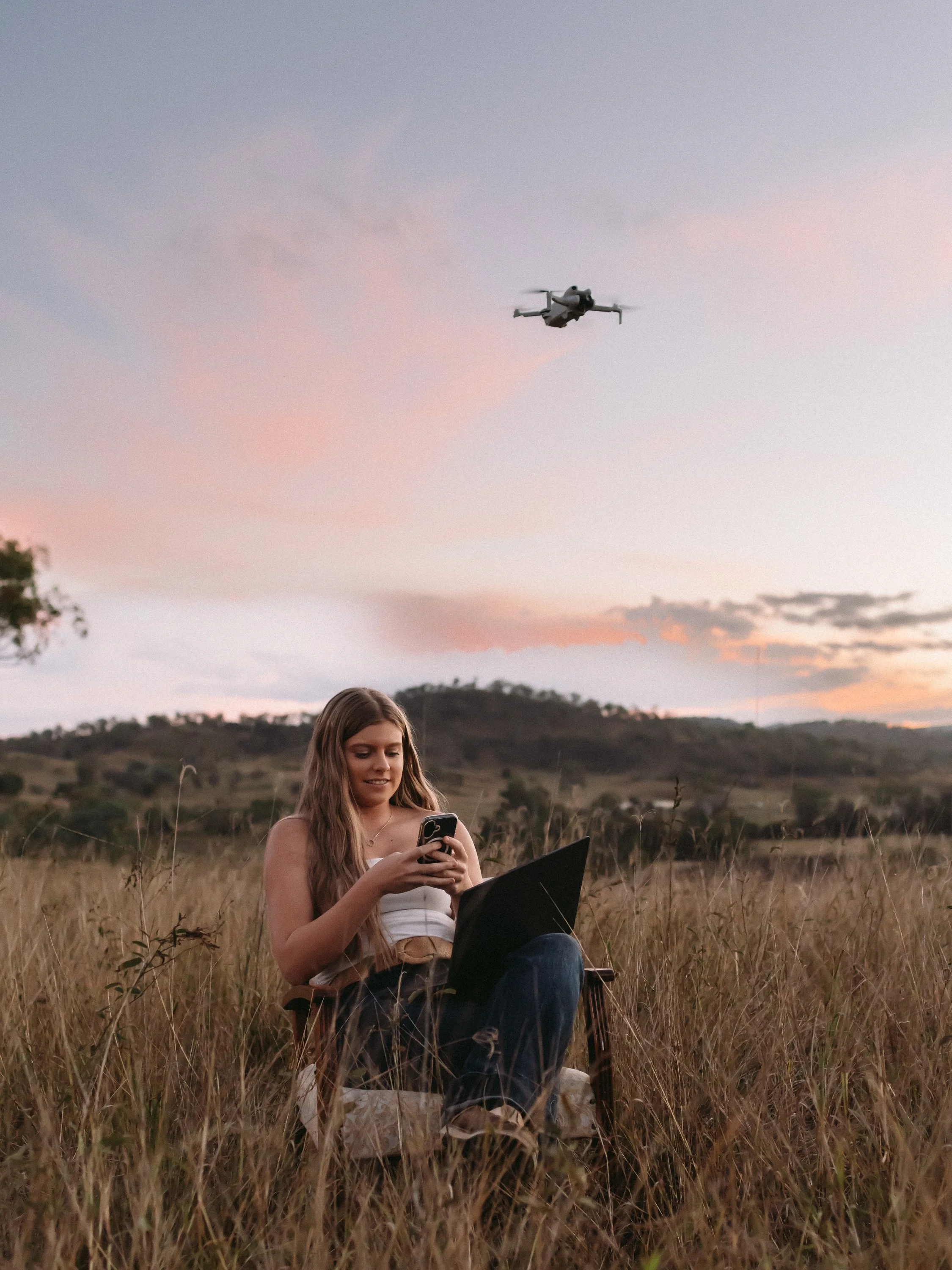 A young woman sitting in a field of tall grass at dusk, using her phone with a drone flying in the sky above her.