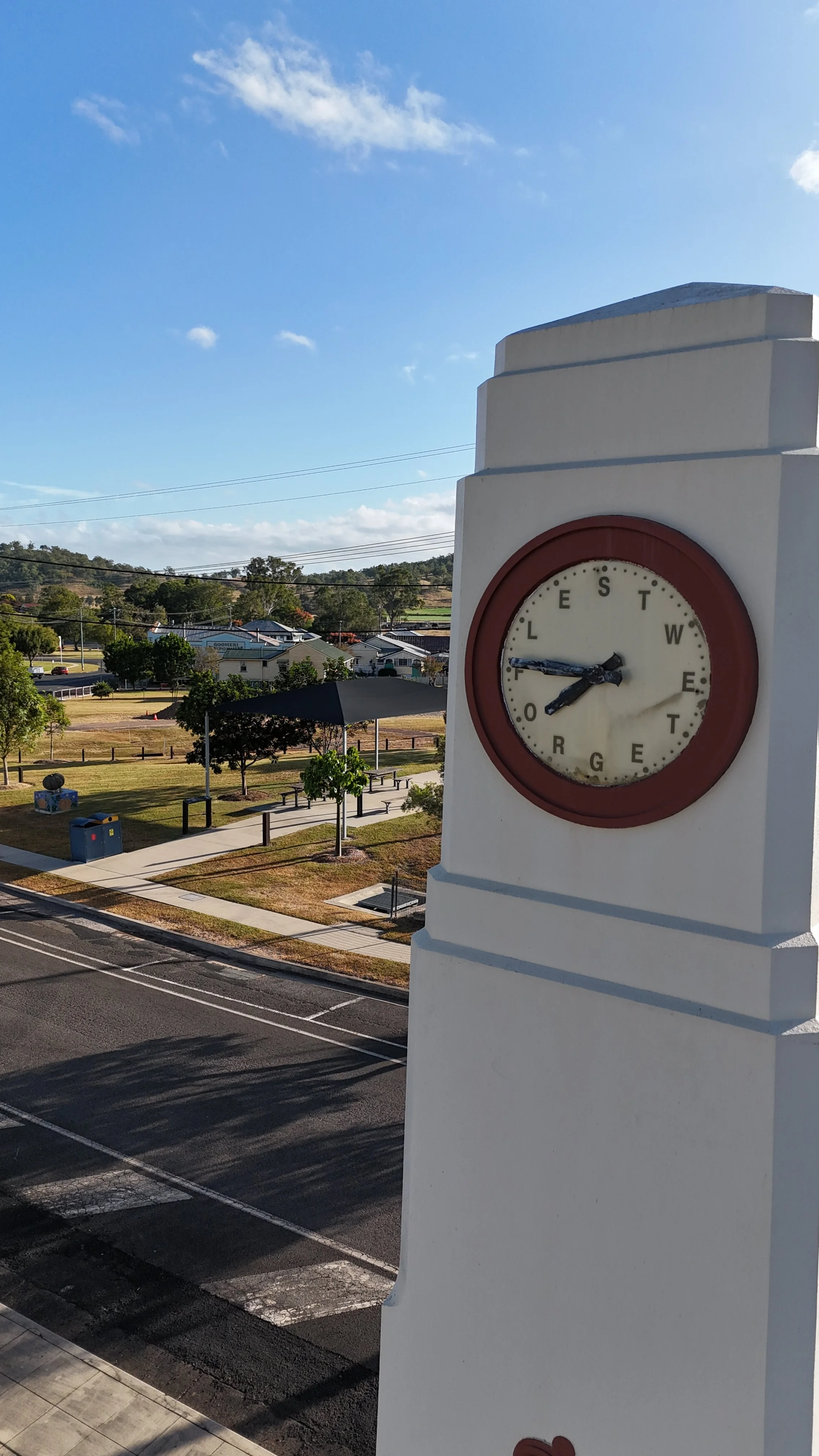 A clock on a white tower showing 2:39, with a park and street below and a blue sky with some clouds in the background.