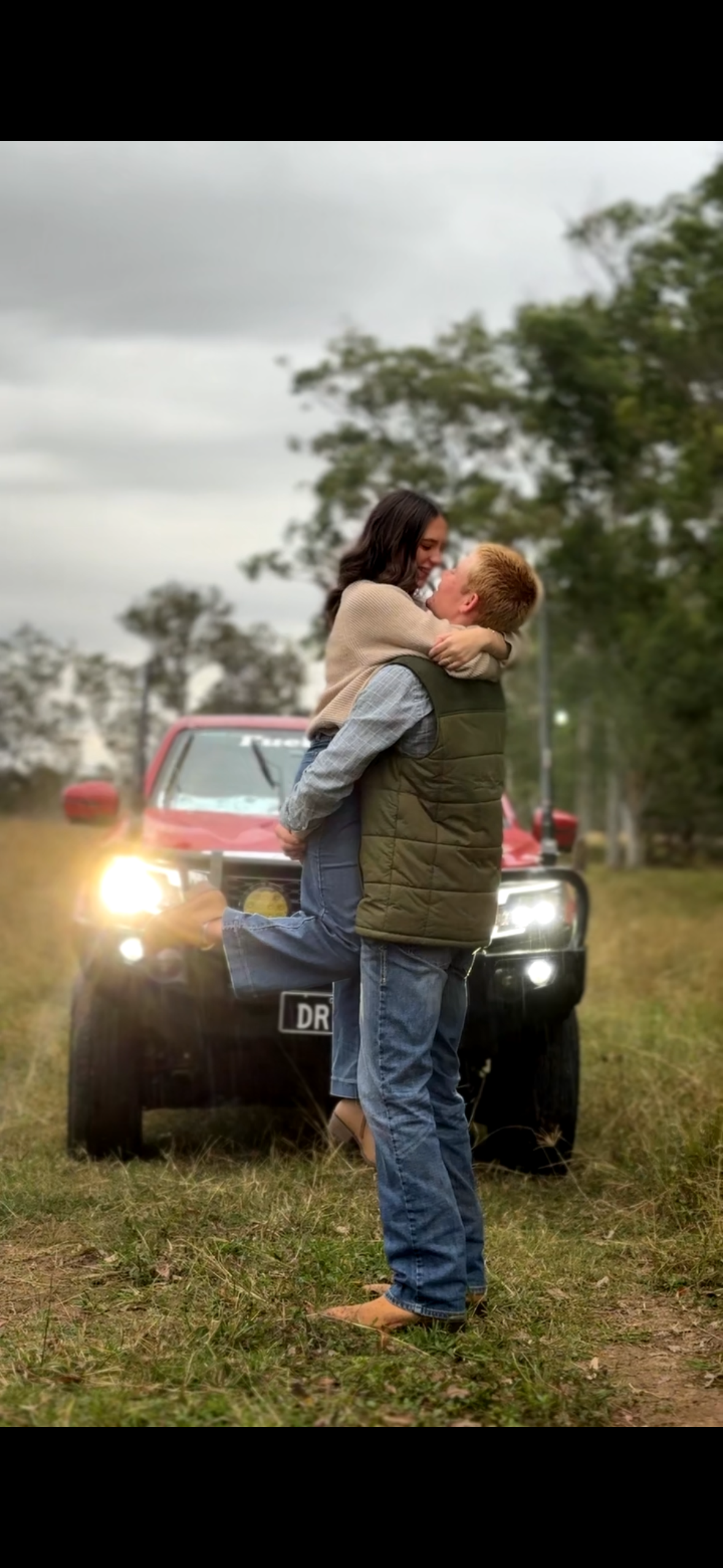 A couple embracing outdoors in front of a red truck, with one person lifting the other and both smiling.