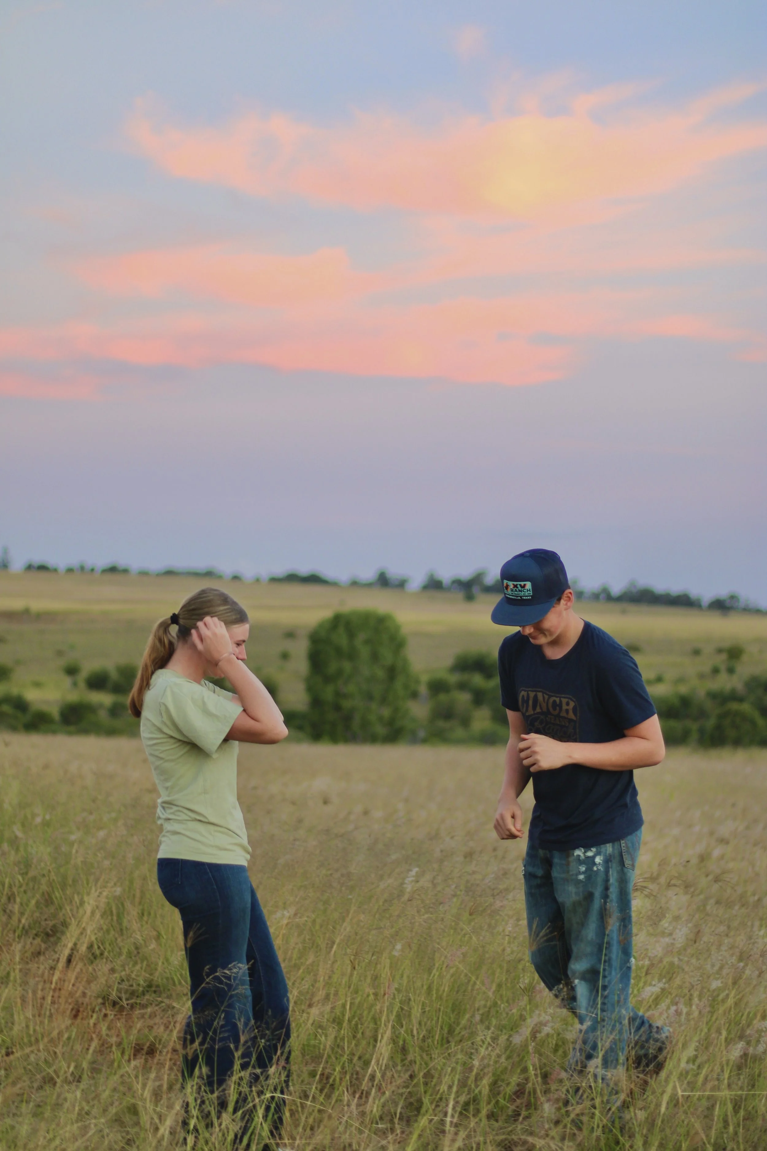 Two children, a girl and a boy, standing in a field of tall grass during sunset. The girl has blonde hair tied in a ponytail and is wearing a light green shirt and jeans. The boy is wearing a black cap, a blue t-shirt, and camouflage pants. They are looking down, with the girl touching her hair and the boy's head slightly bent.