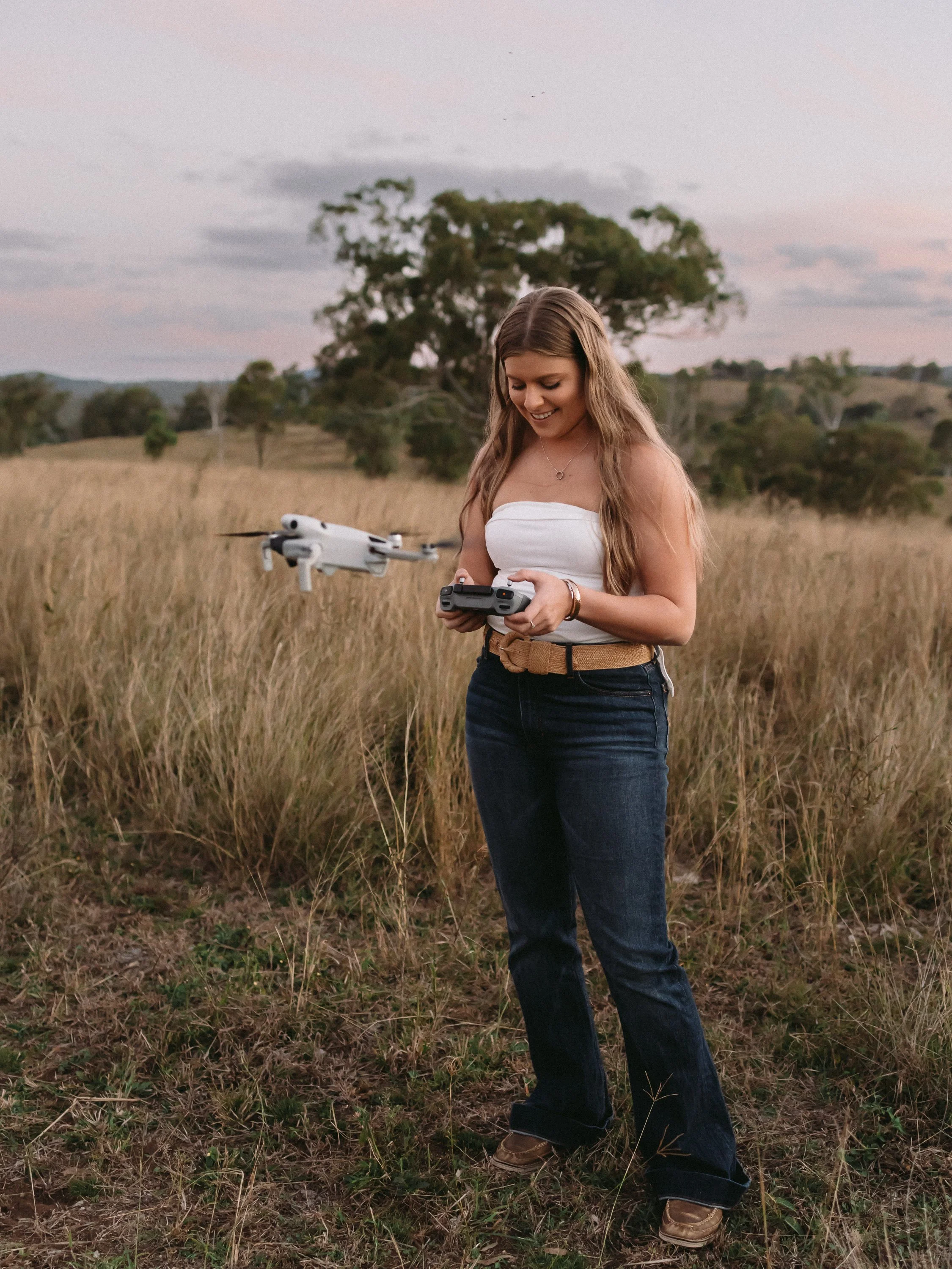 A young woman with long wavy hair wearing a white strapless top, dark jeans, and brown shoes, holding a drone controller while standing in a grassy field with trees and a cloudy sky in the background.