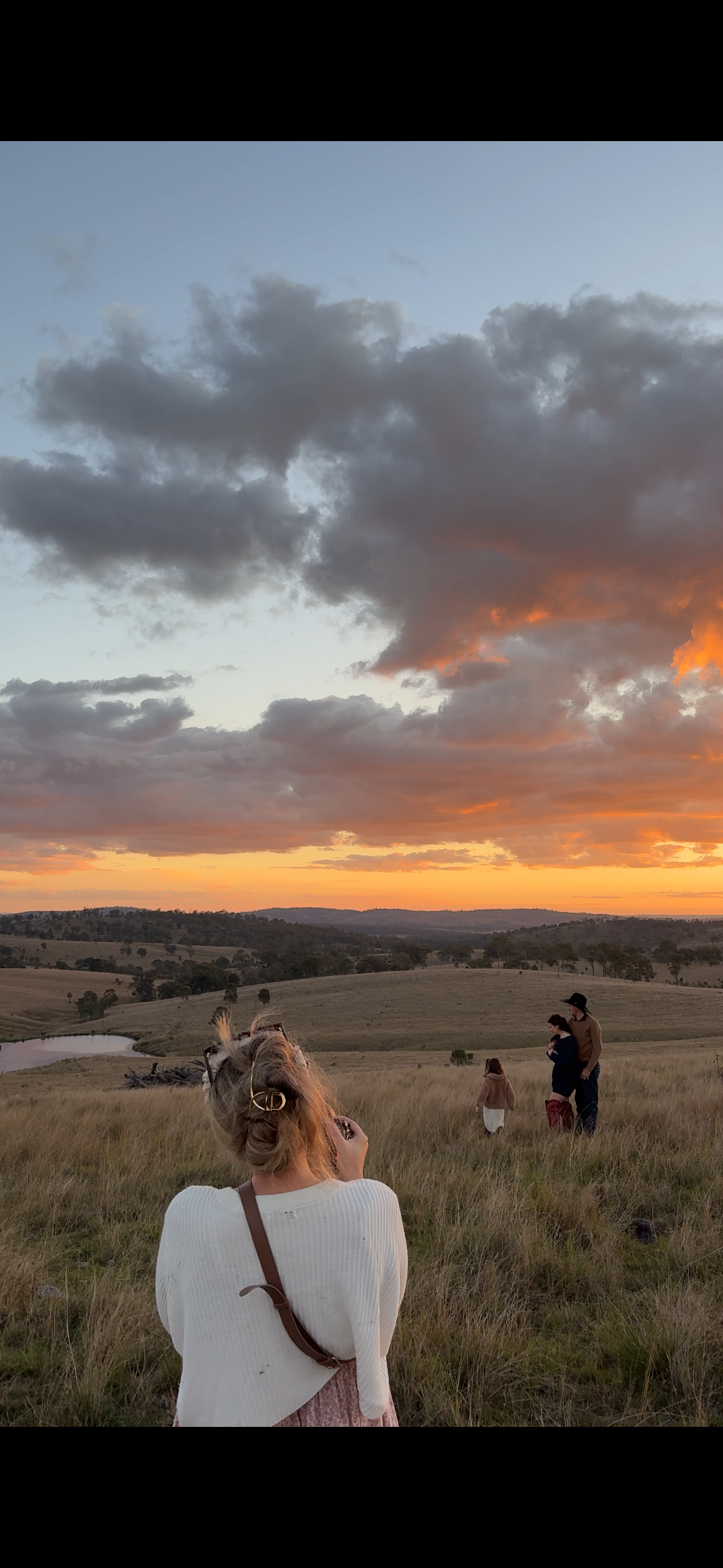 People enjoying a sunset landscape with rolling hills, a pond, and a dramatic sky with dark clouds and orange hues.