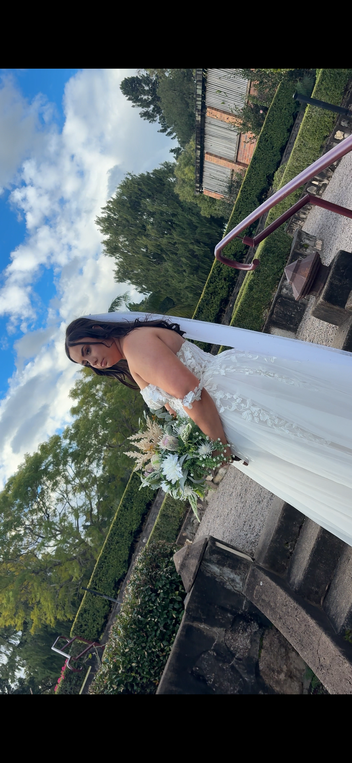 A bride in a white wedding dress holding a bouquet of flowers outdoors on a stone pathway with trees and a fence in the background.
