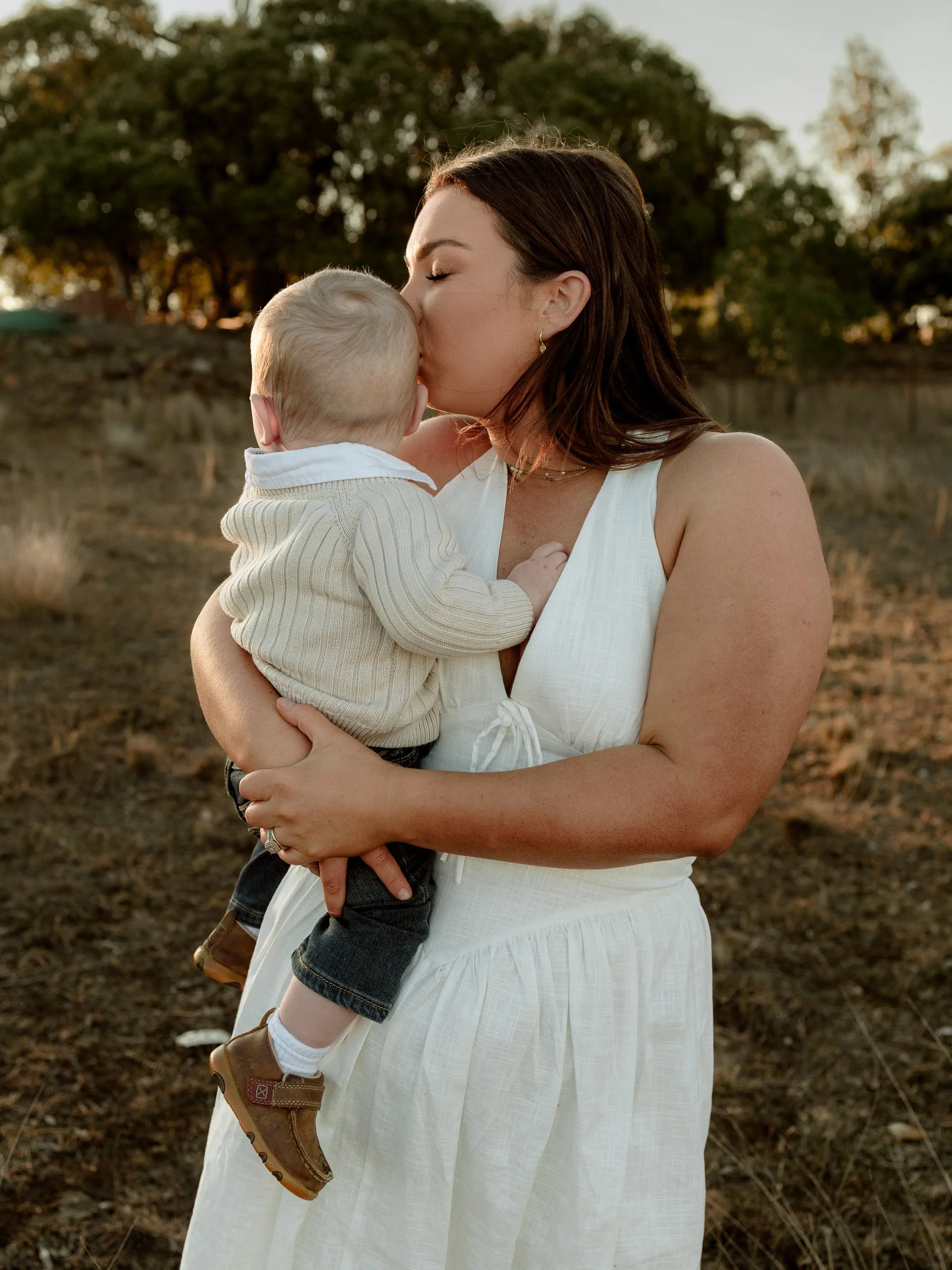 A woman in a white dress holding and kissing a small child outdoors, with trees and a stone wall in the background during sunset or late afternoon.
