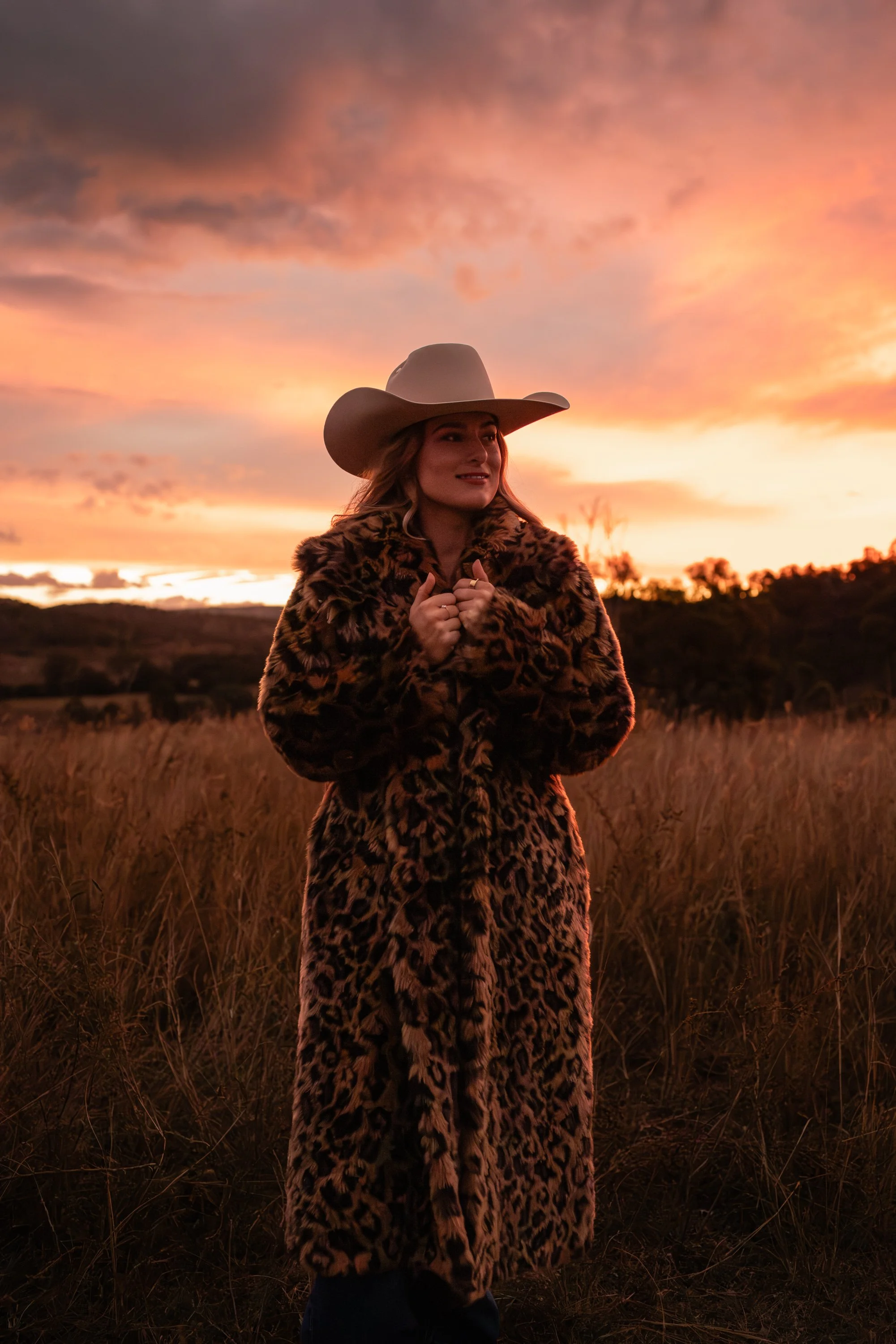 A woman standing in a field at sunset, wearing a leopard print fur coat and a wide-brimmed hat, with her hands clasped together.