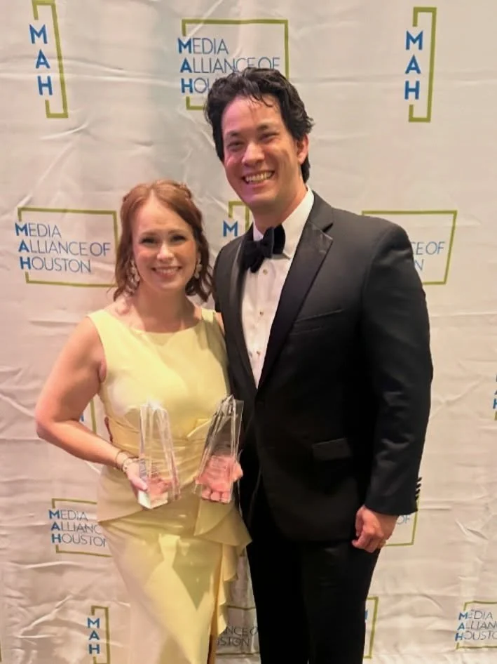 A woman and a man in formal attire smiling at an event with a backdrop that reads 'Media Alliance of Houston'; the woman is holding glass awards.