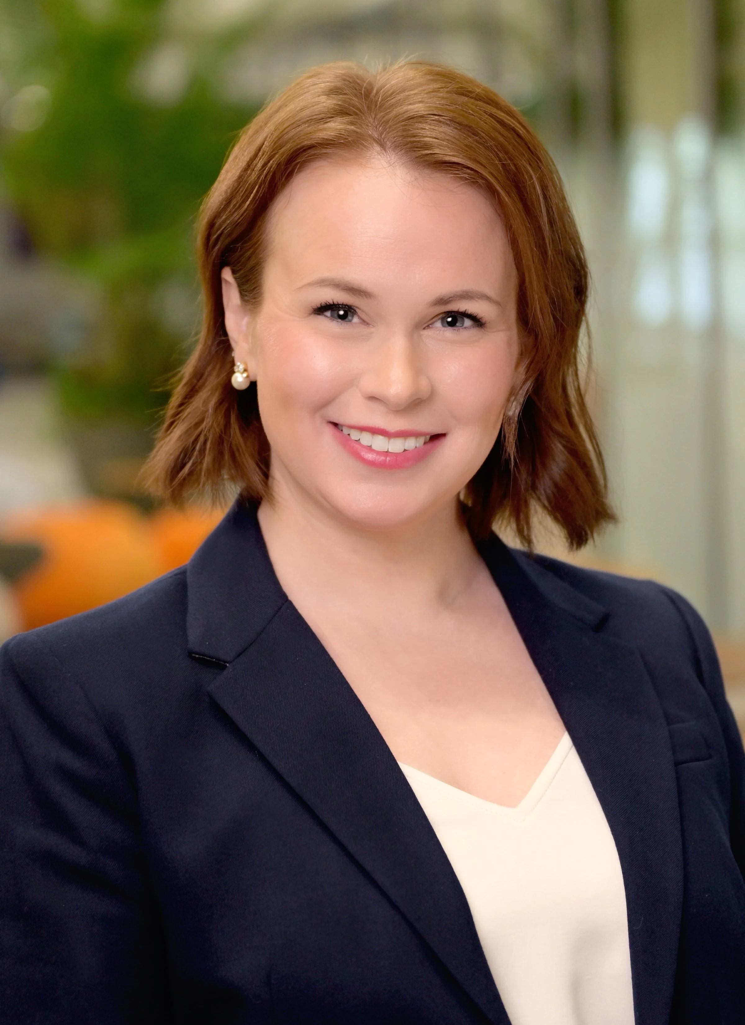 Close-up of a smiling woman with shoulder-length red hair, wearing pearl earrings, a dark blazer, and a white V-neck top, with a blurred background of green and orange foliage.
