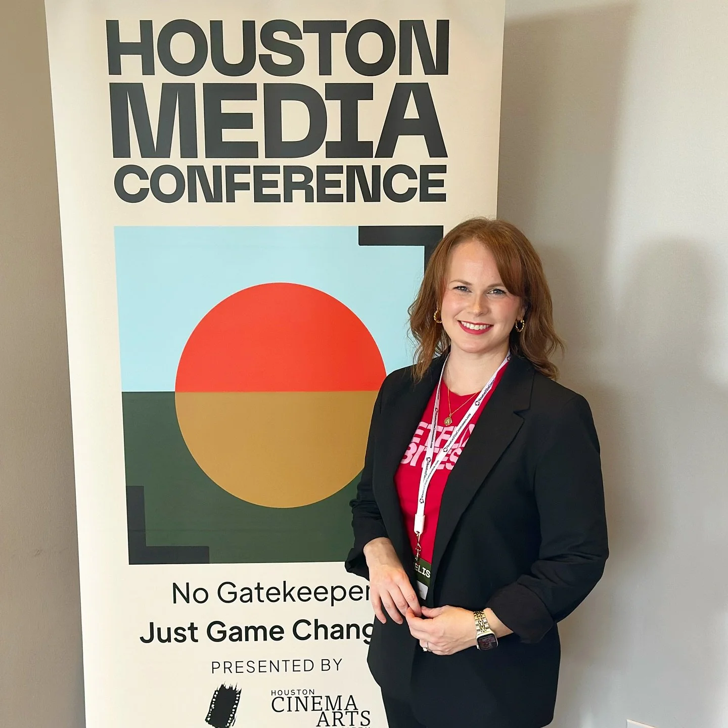 A woman with brown hair dressed in a black blazer and red top standing next to a Houston Media Conference sign, smiling for the camera.