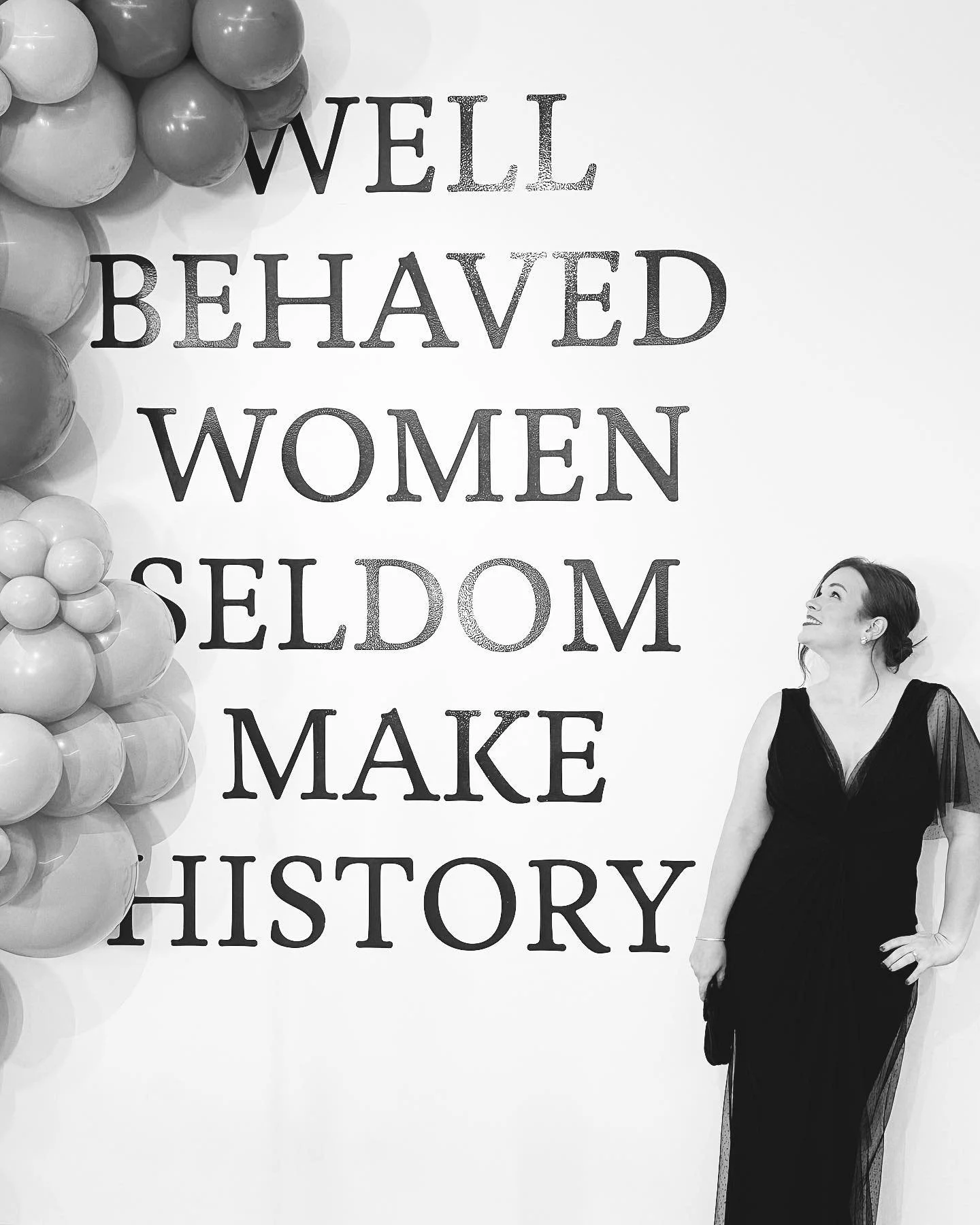 A woman in a black evening gown stands next to a wall decorated with black and white balloons and a quote that reads: 'WELL BEHAVED WOMEN SELDOM MAKE HISTORY'.