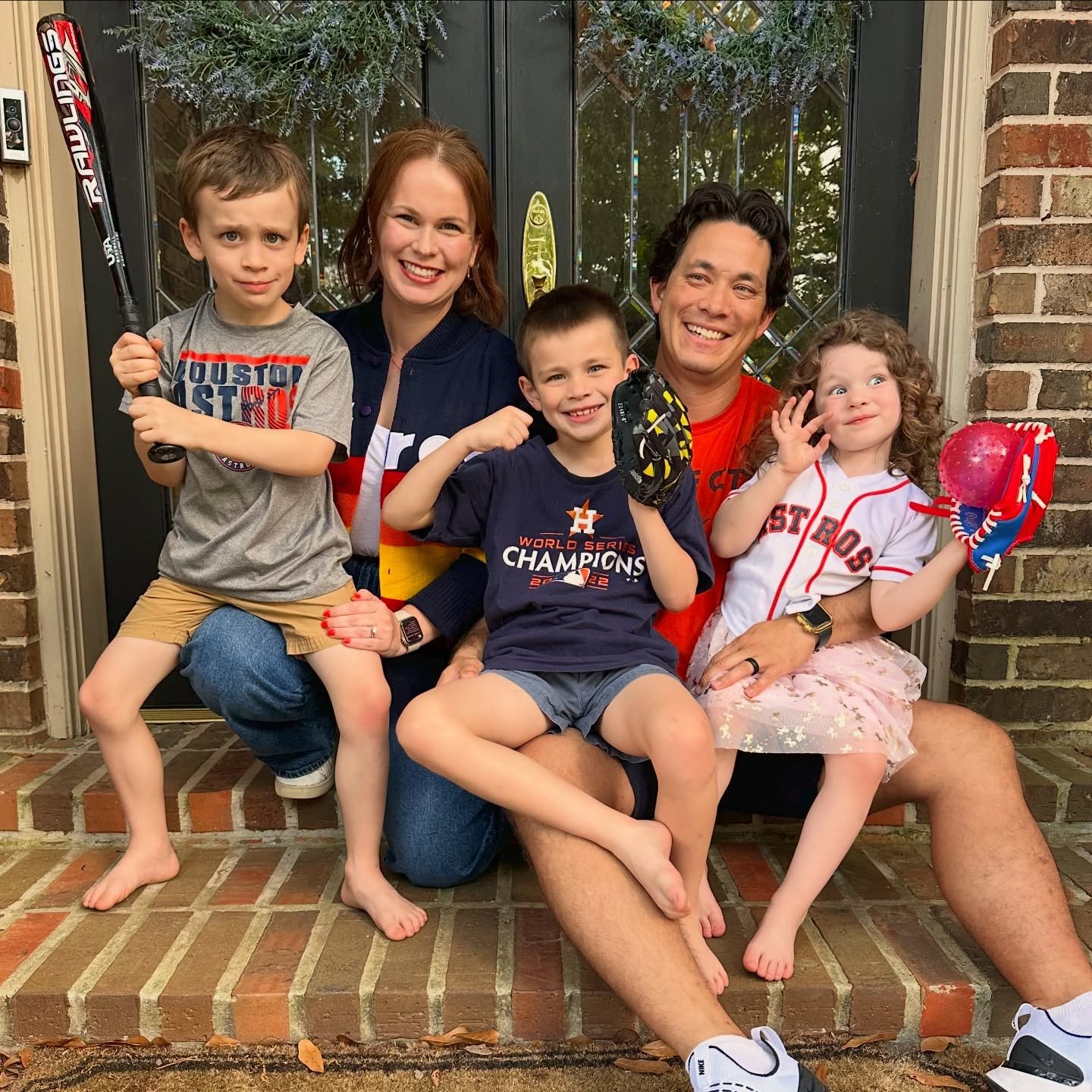 A family of five poses together on the front porch of a brick house, dressed in Houston Astros baseball team apparel, holding baseball gear, and smiling.