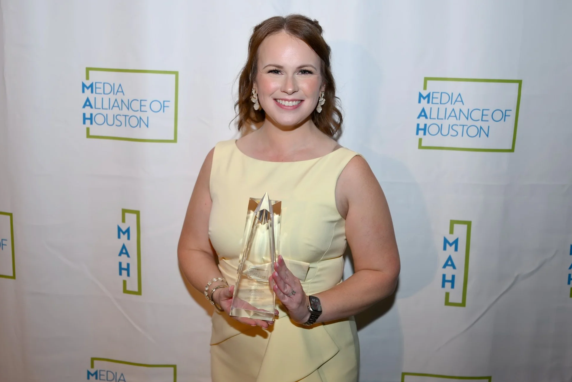 Woman in yellow dress holding an award and smiling at a Media Alliance of Houston event.