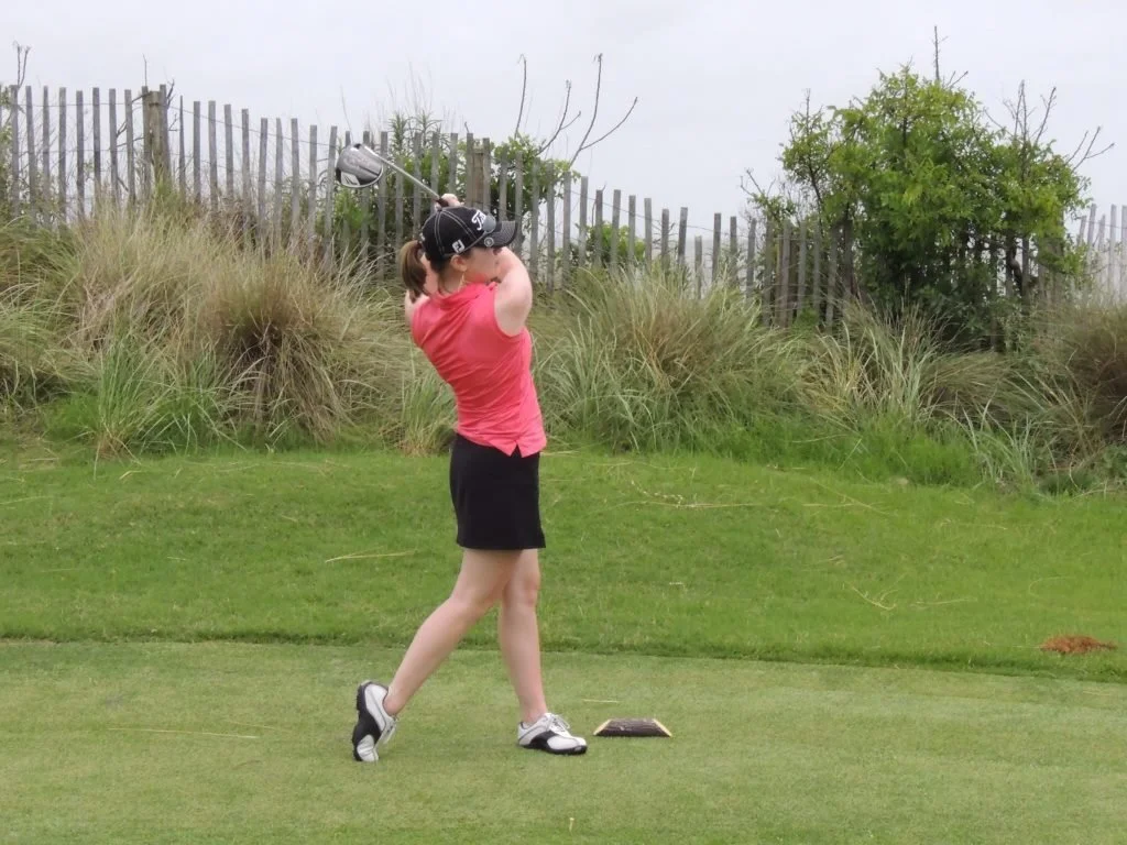 A girl in a pink shirt and black skort swinging a golf club on a golf course.