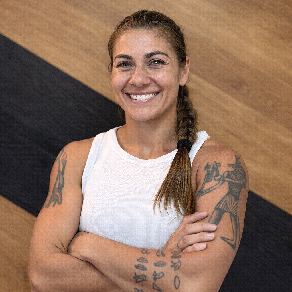 Smiling woman with tattoos on her arms, wearing a white sleeveless top, standing with arms crossed against a wooden and black textured background.