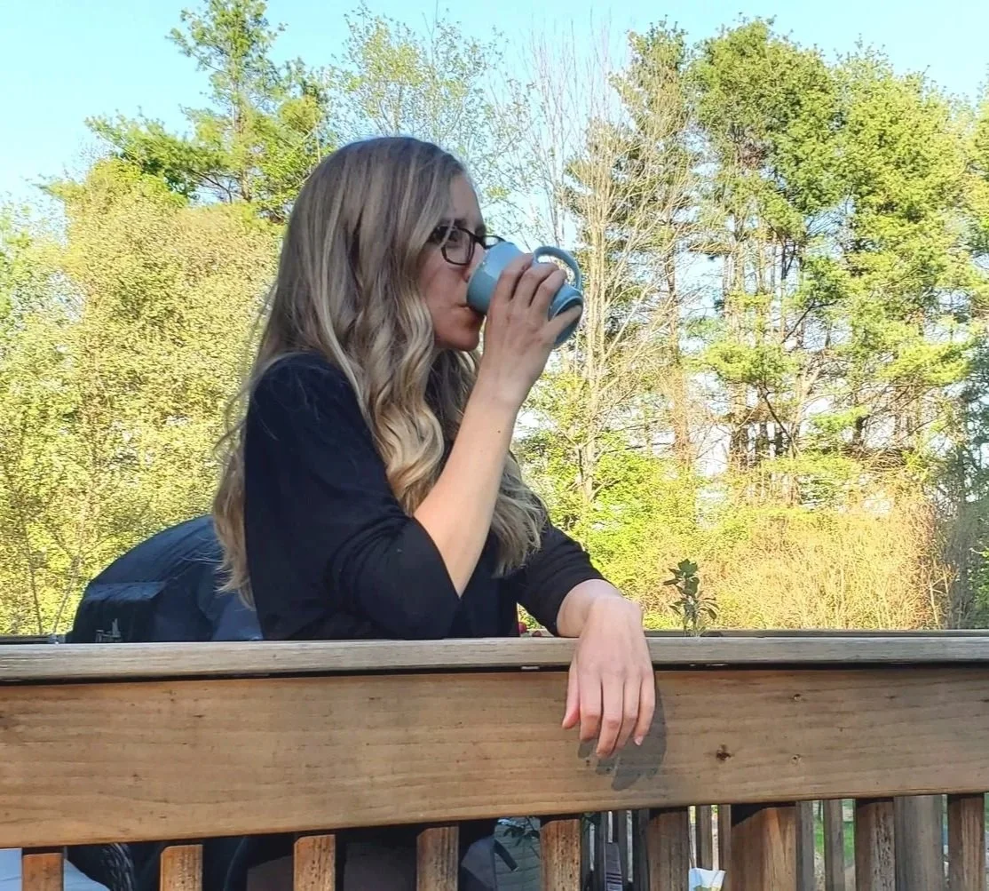 A young woman with long wavy blonde hair, wearing glasses, sits outdoors and drinks from a blue mug. She is leaning on a wooden railing with trees and a clear sky in the background.