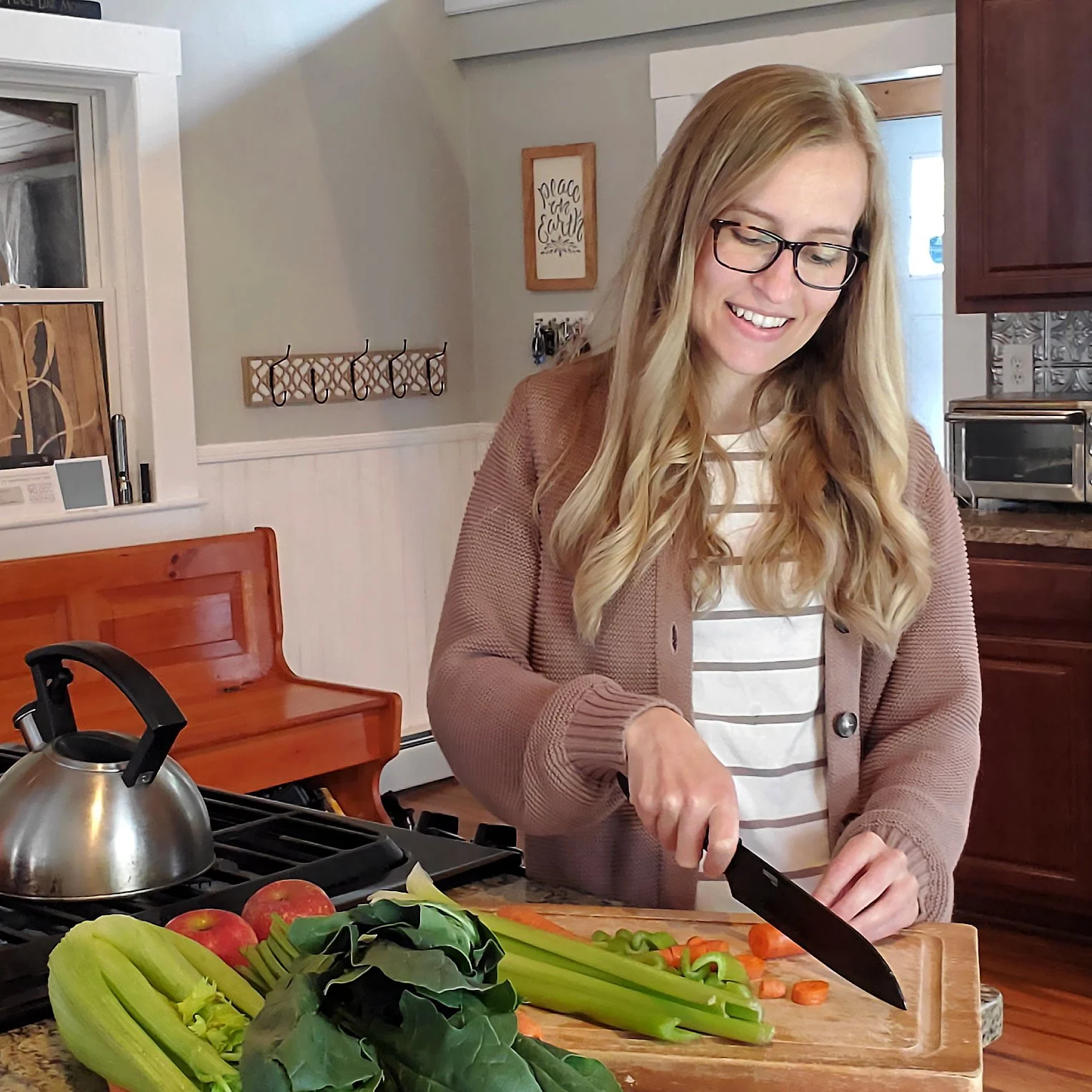 A woman with glasses is chopping vegetables, including carrots and celery, on a wooden cutting board in a kitchen with apples and leafy greens in the foreground.
