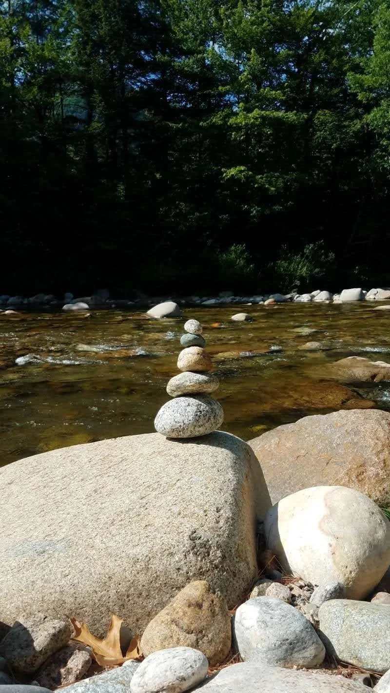 5 small rocks balancing on top of each other, with a river in the background of the image..