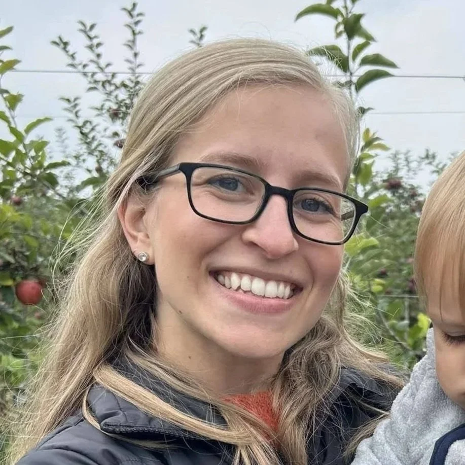 A woman with blonde hair, glasses, and a black jacket, smiling outdoors, surrounded by apple trees.