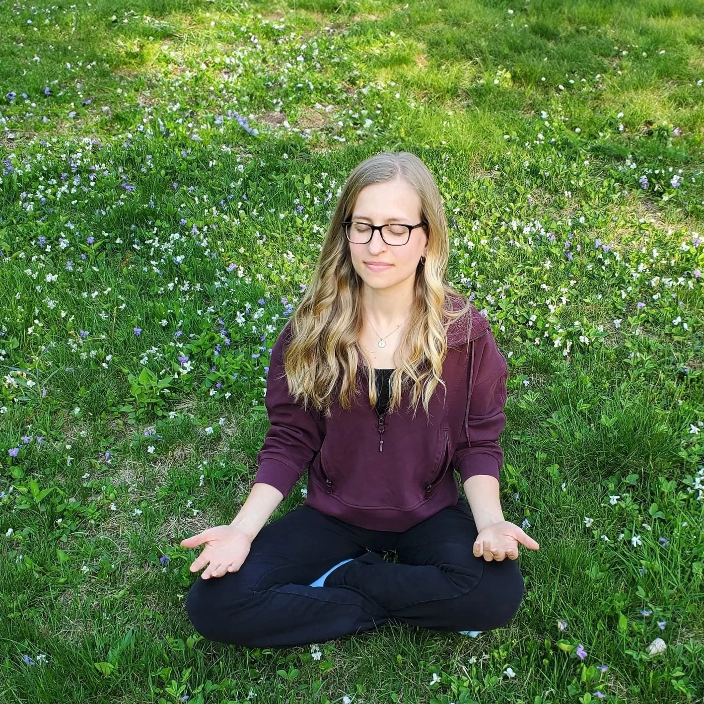 A young woman with glasses, wearing a maroon jacket, sitting cross-legged on a grassy field with small purple and white flowers, meditating with eyes closed and hands resting on knees.