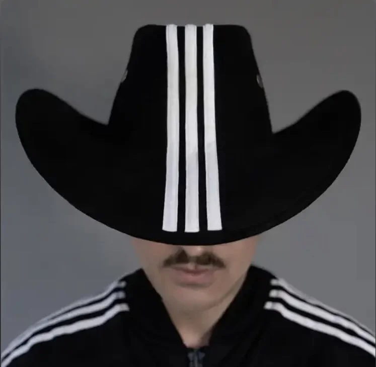 Person wearing a large black hat with white stripes and a black Adidas tracksuit with white stripes, against a gray background.