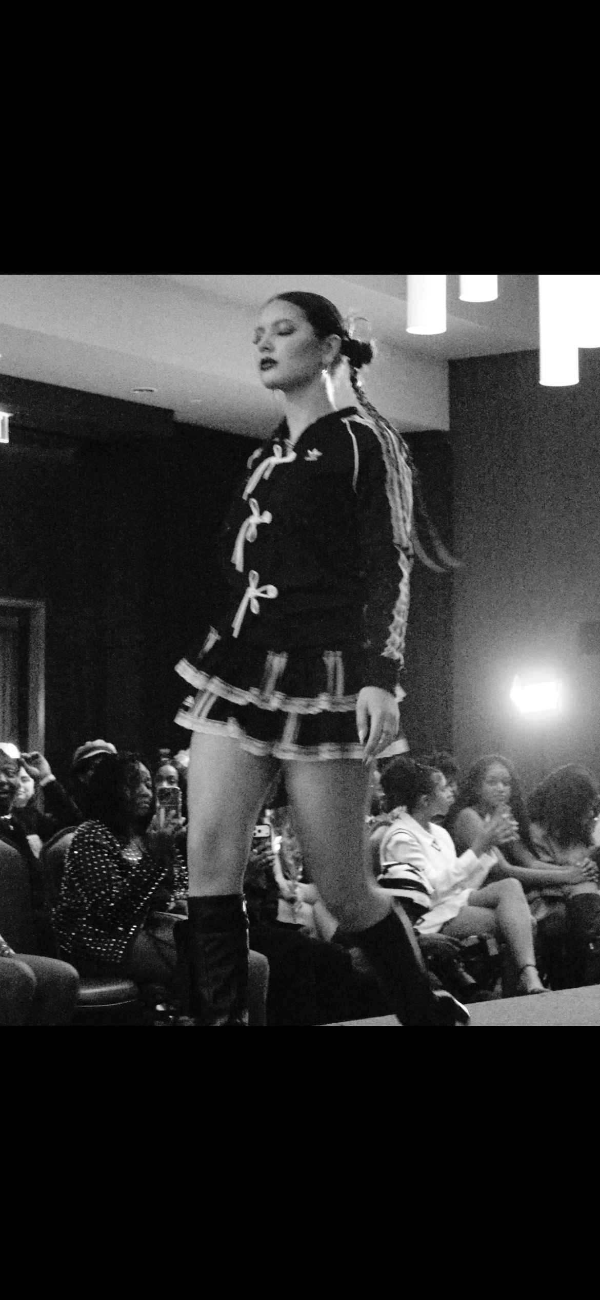 A woman walking on a runway during a fashion show, with an audience watching and taking photos.