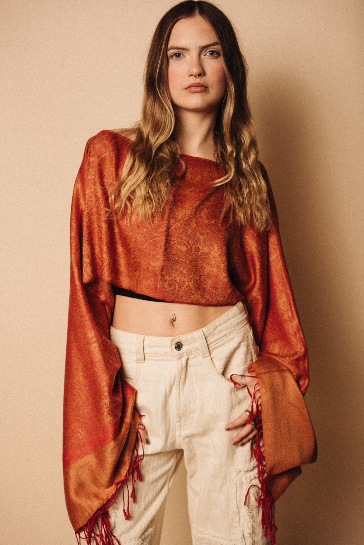 A young woman with long wavy hair, wearing a cropped patterned orange top, beige high-waisted pants, and a long orange shawl with red fringe details, standing against a beige background.
