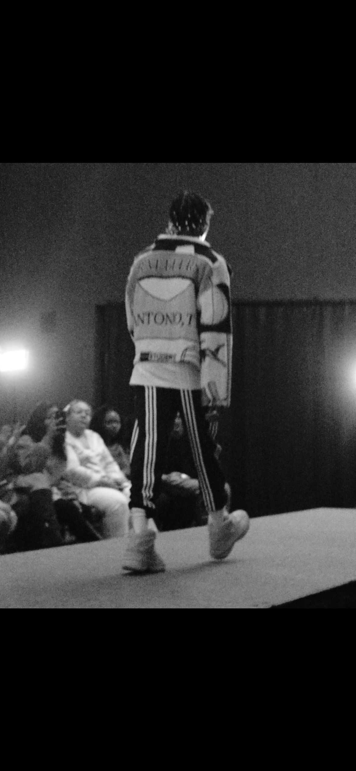 A person walking on stage in front of an audience, seen from behind, wearing a jacket, striped pants, and sneakers, with an audience watching.