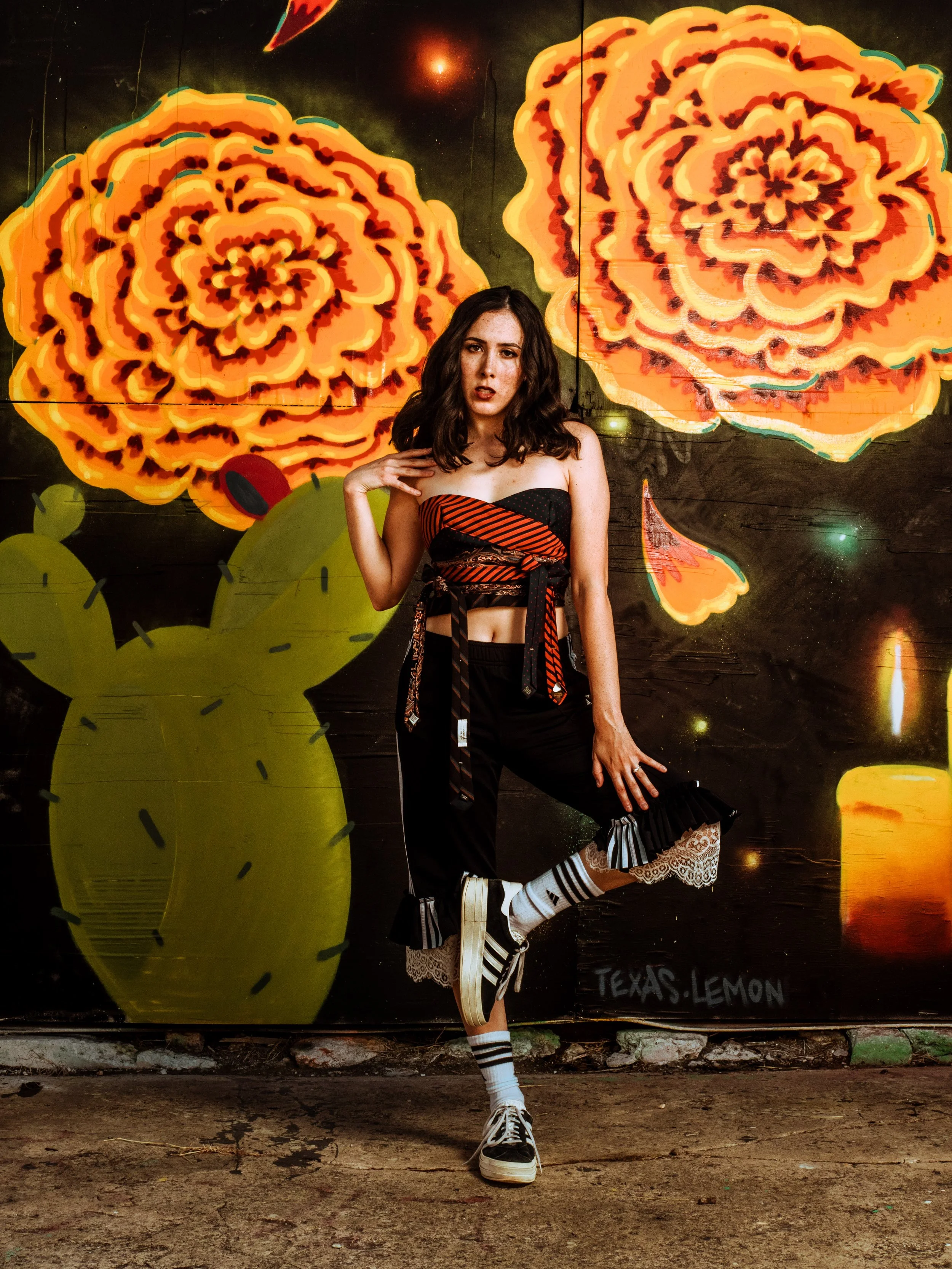 A young woman with dark wavy hair and freckles standing in front of a colorful mural of large yellow Texas lemons. She is dressed in a black and red striped top, black pants with ruffled hems, and white sneakers. She is wearing striped socks, has one