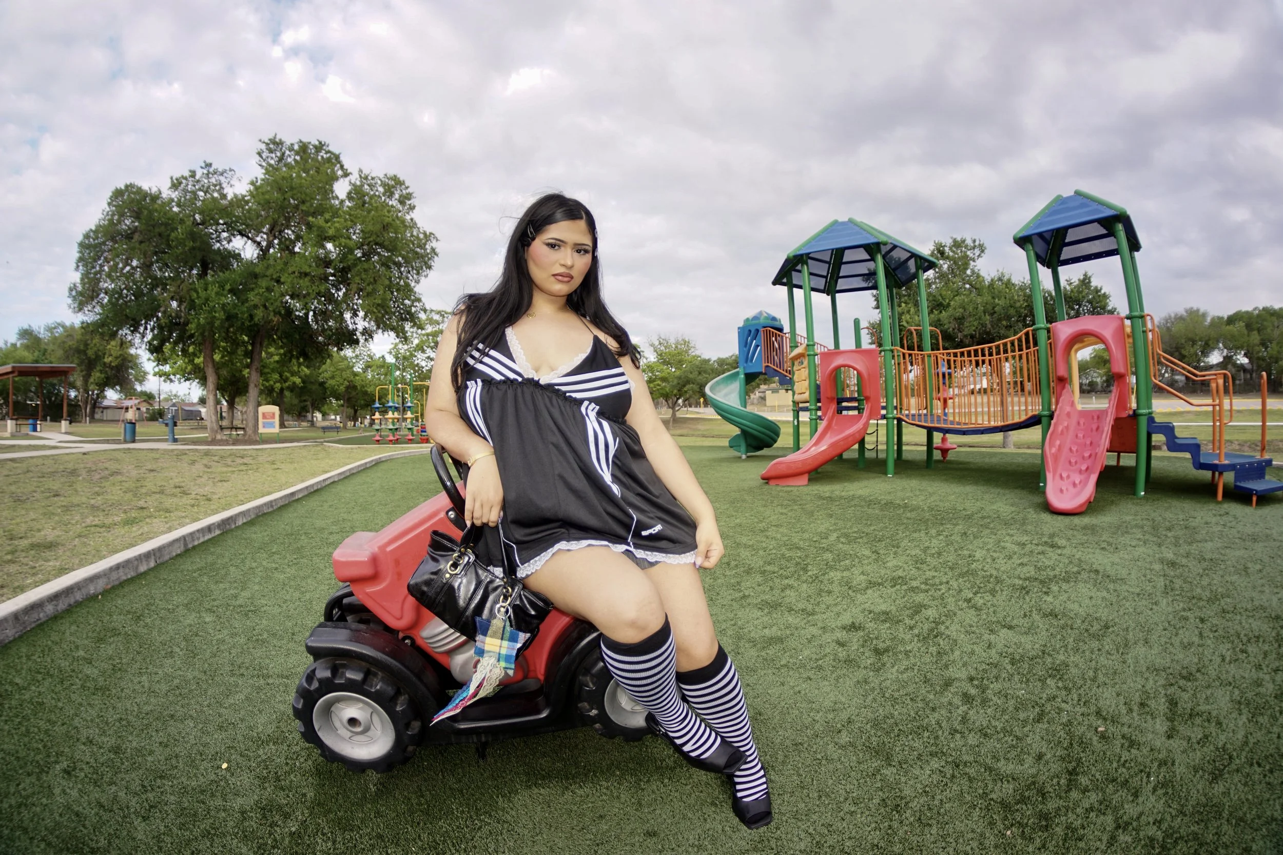 Young woman in a black and white striped dress and knee-high socks sitting on a toy car in a park with playground equipment and trees.