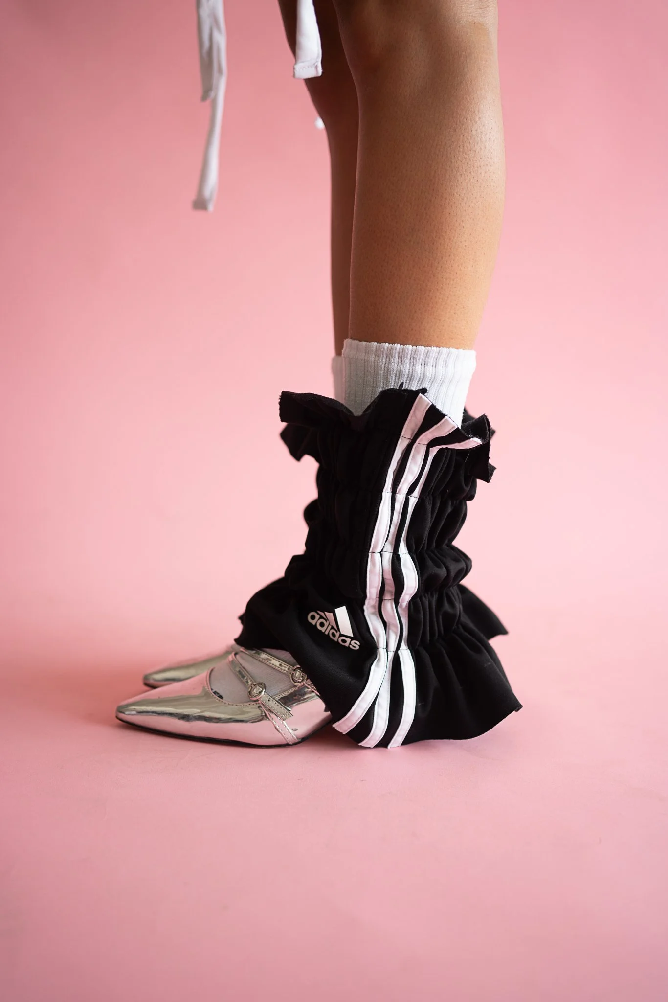 Close-up of a person crouching on pink background, wearing metallic silver pointed shoes, black Adidas track pants with white stripes, white socks, and part of a white garment visible.