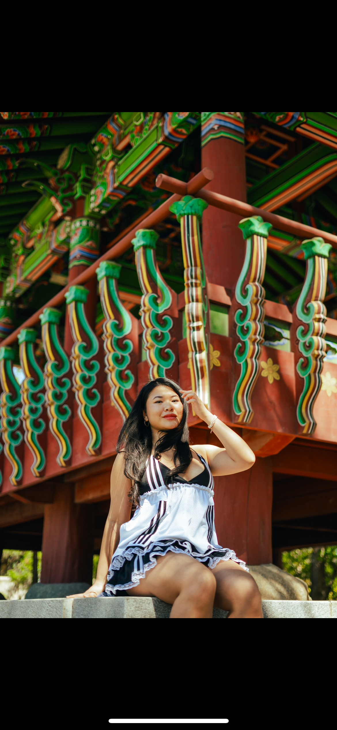 A woman sitting outdoors on a stone surface in front of a colorful, traditional Asian-style structure with intricate woodwork and decorative details.