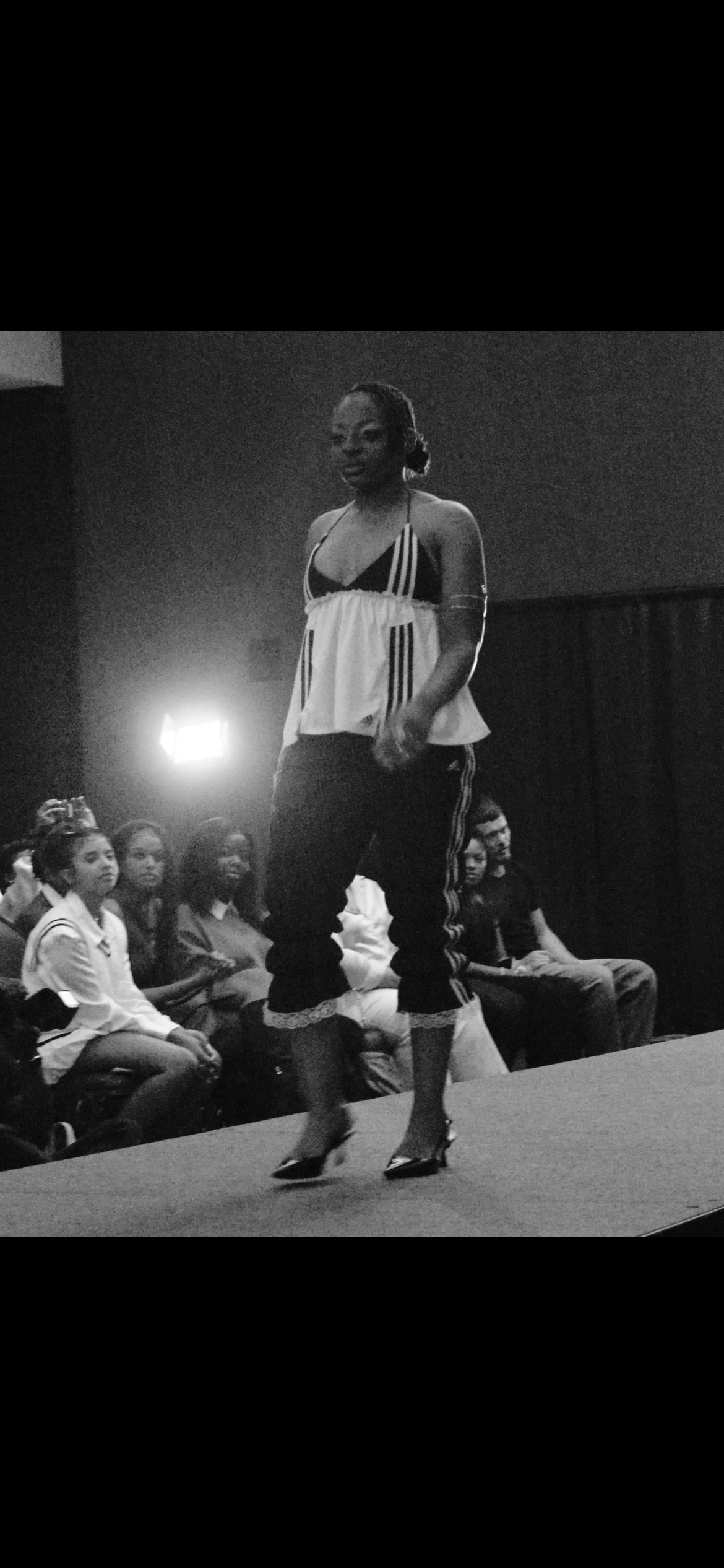 A woman walking on a runway at a fashion show, wearing a loose tank top and track pants, with a group of seated audience members watching.