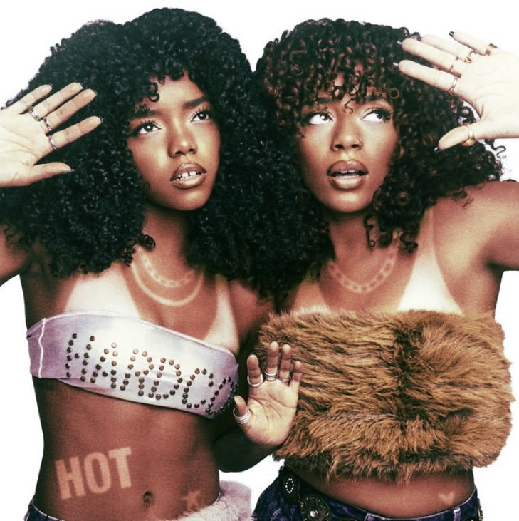 Two women with curly hair, posing with their hands near their heads. One woman is wearing a strapless top with 'VANROCK' written on it, and a temporary tattoo that says 'HOT' on her stomach. The other woman is wearing a fur top. Both are looking slightly upward and to the side.