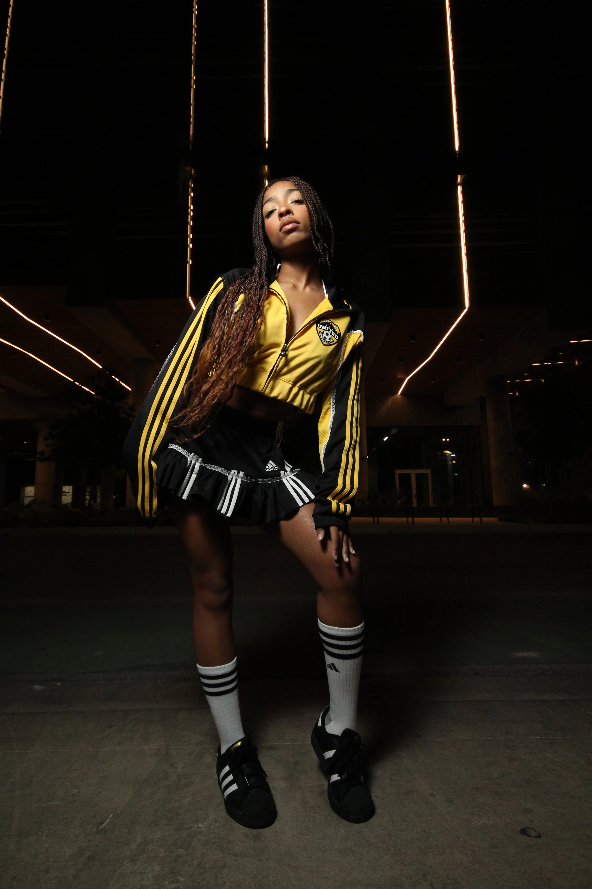 A young woman standing outdoors at night, wearing a black and yellow Adidas jacket, a black skirt with white stripes, knee-high white socks with black stripes, and black sneakers. She has long braided hair and a confident pose, with neon lights in th