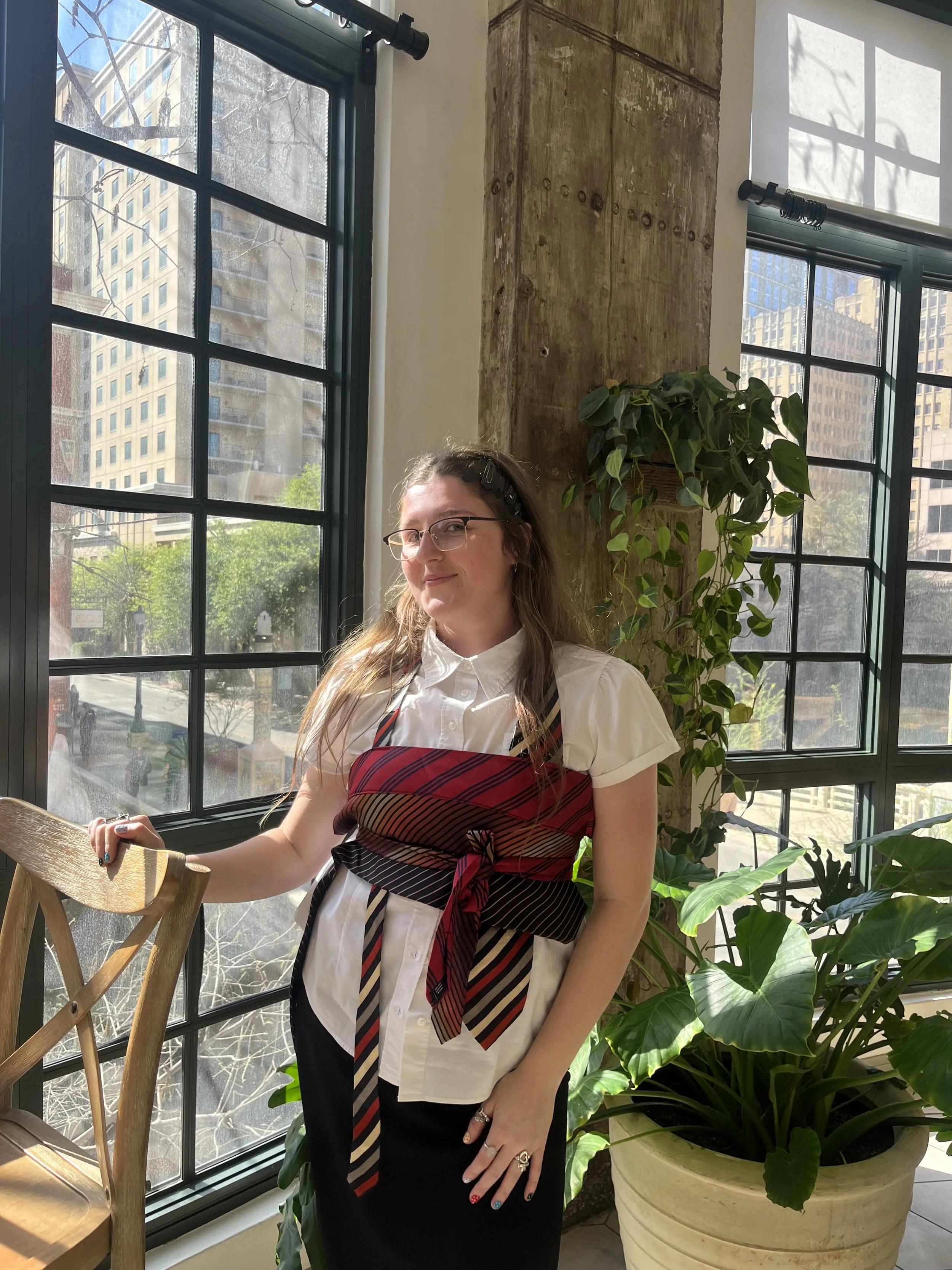 A woman wearing glasses, a white shirt, and a black skirt, standing indoors by large windows with city buildings outside. She is holding a wooden chair and has colorful nail polish and rings on her fingers. There are green plants in a large beige pot and a small potted plant on a wooden surface.