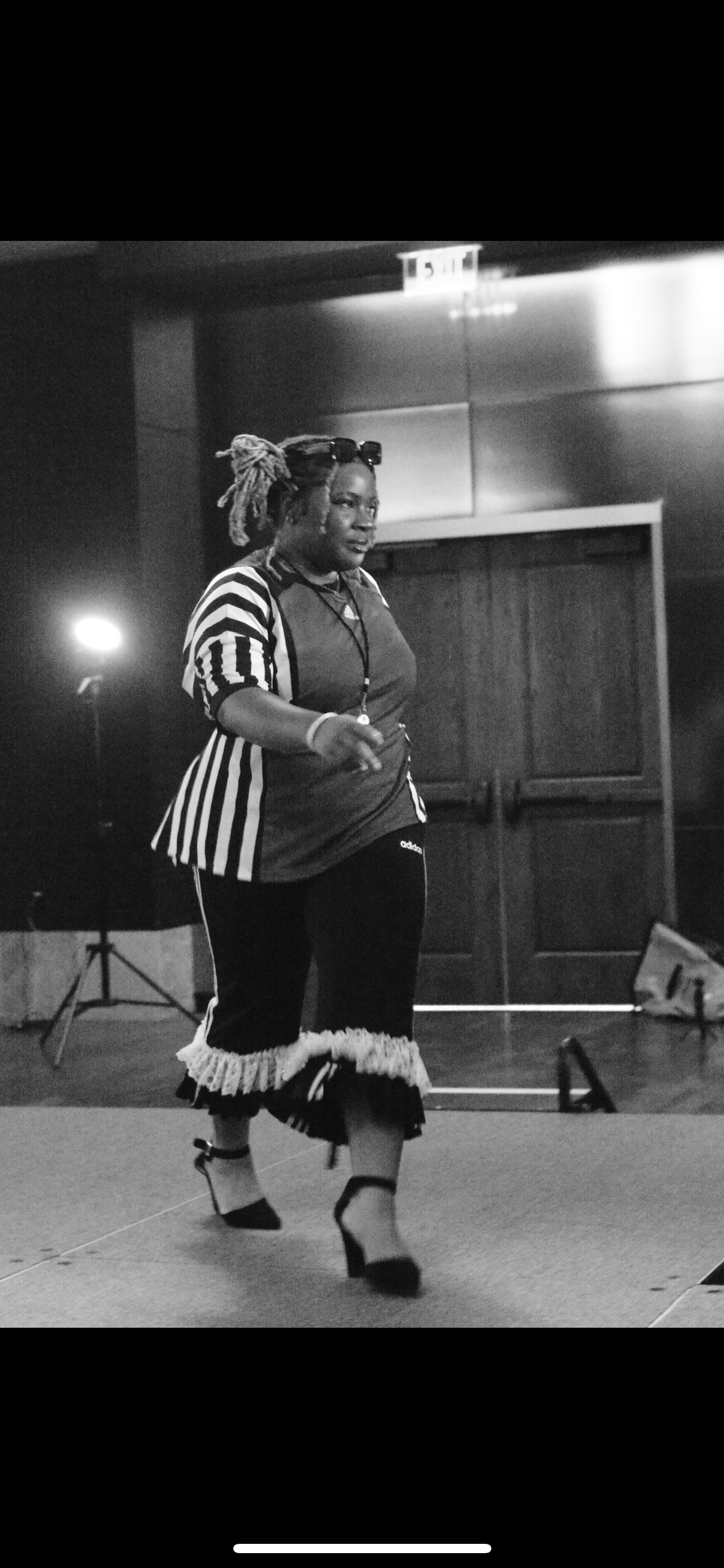 A woman with dreadlocks wearing a striped referee shirt, decorative skirt, and high heels, dancing or performing on a stage in an indoor setting.