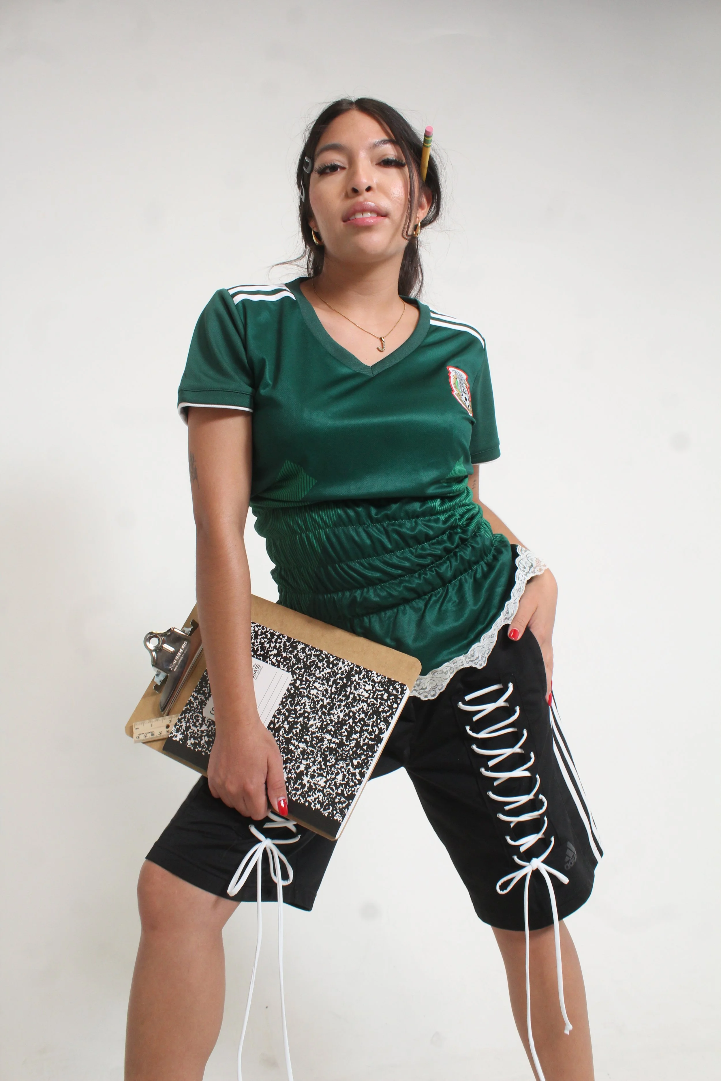 A young woman wearing a green sports jersey and black shorts with white laces, holding a clipboard and standing against a plain white background.