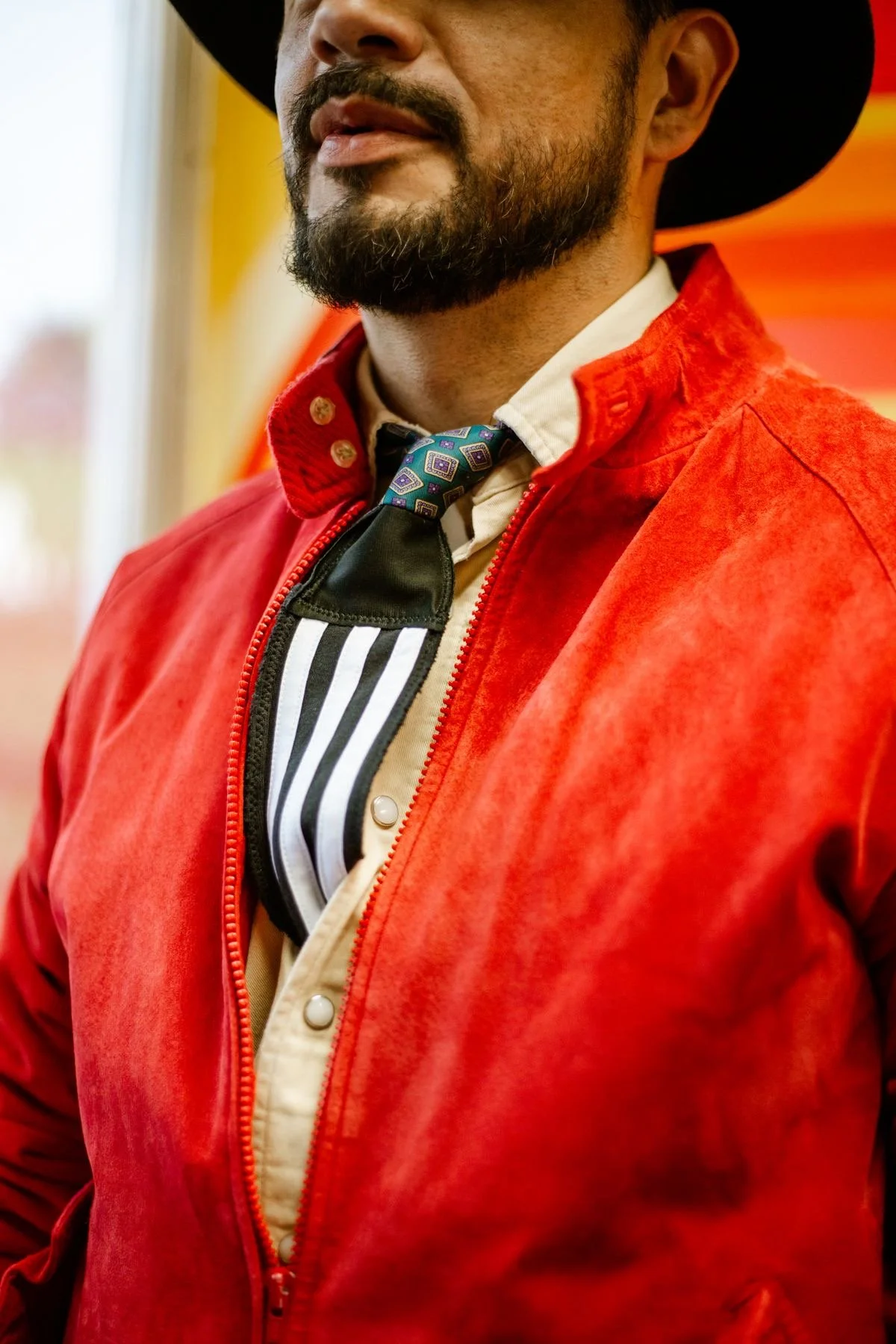 Close-up of a man wearing a black hat, red jacket, and layered clothing with a striped shirt, patterned tie, and beige shirt underneath. The background is blurred with warm colors.