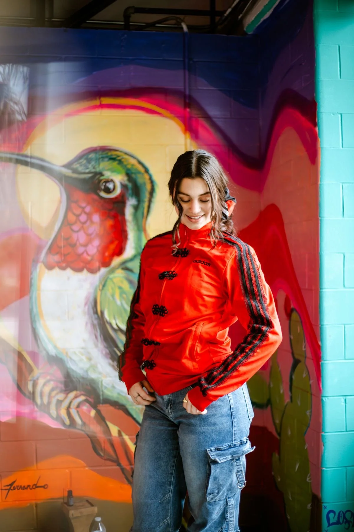 A young woman with dark hair in a braid, wearing a red Adidas jacket and blue cargo jeans, standing in front of a colorful graffiti mural of a hummingbird on a brick wall.