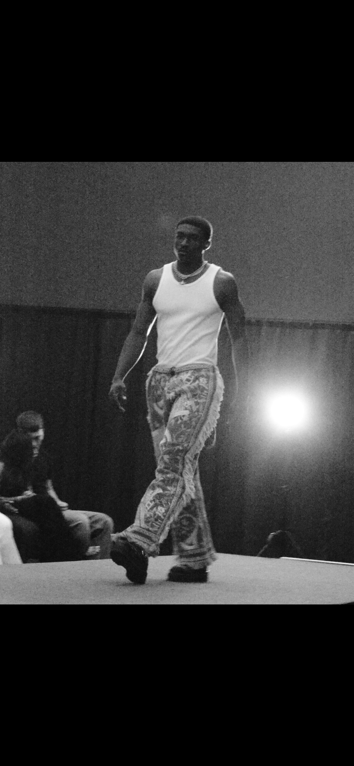 Black and white photo of a young man walking on a stage, wearing a tank top, patterned pants, and a necklace, with a bright light in the background.