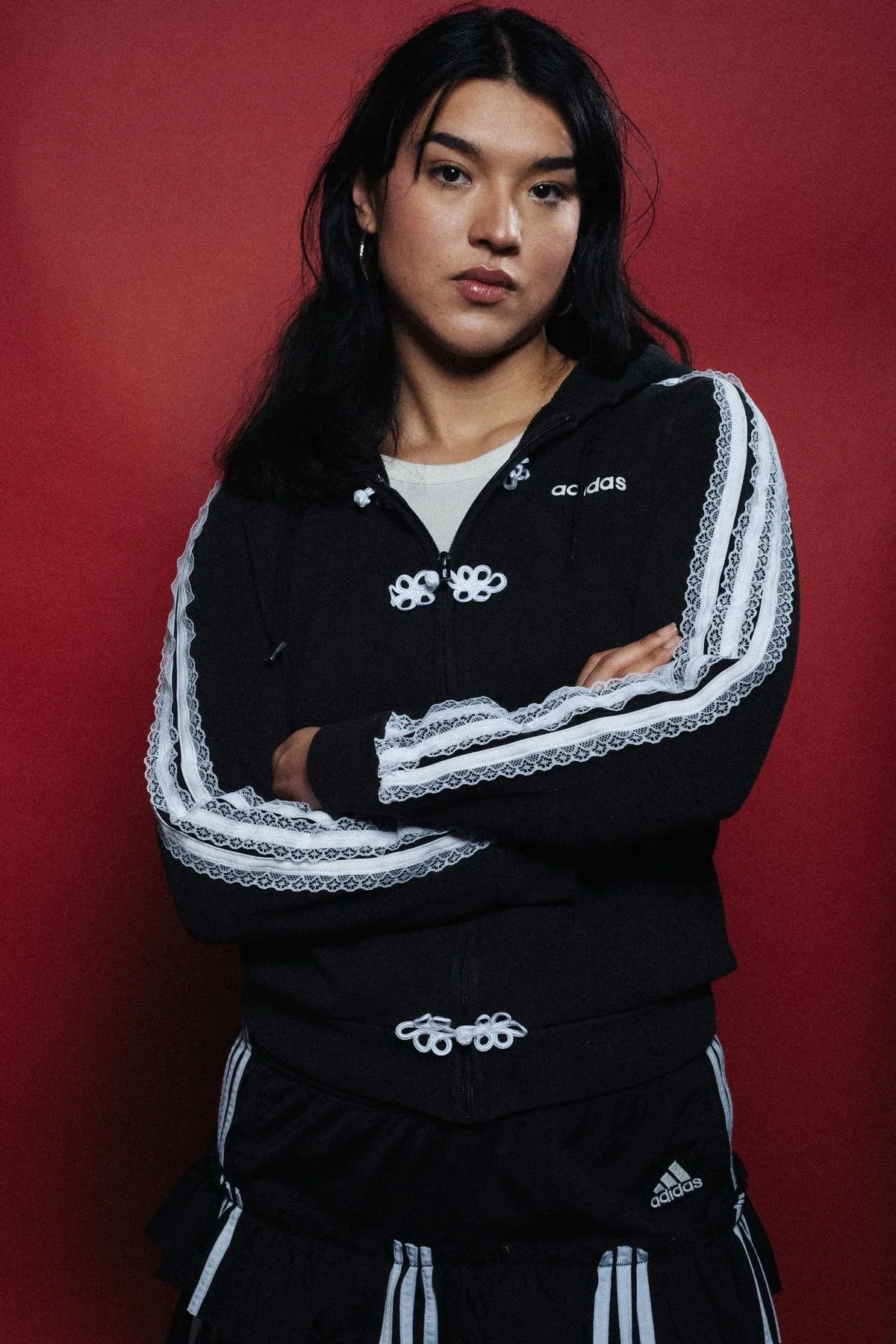 A young woman with dark hair and hoop earrings, wearing a black Adidas hoodie with white stripe details and lace accents, standing against a solid red background with her arms crossed.