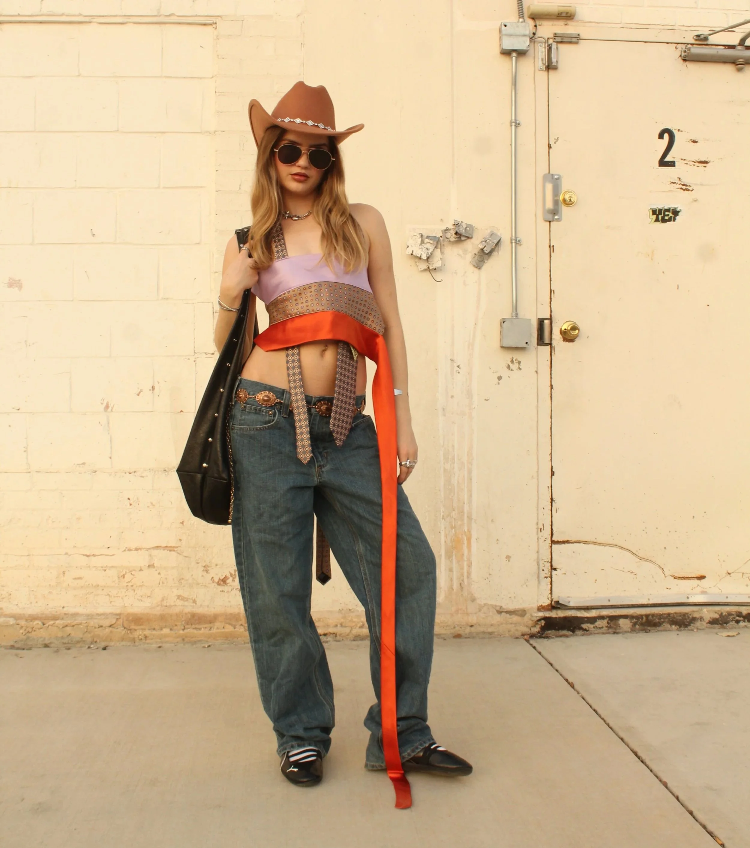A young woman with long wavy hair wearing sunglasses, a cowboy hat, a multi-colored crop top, high-waisted jeans, and black sneakers, standing against a beige brick wall with an industrial door beside her.