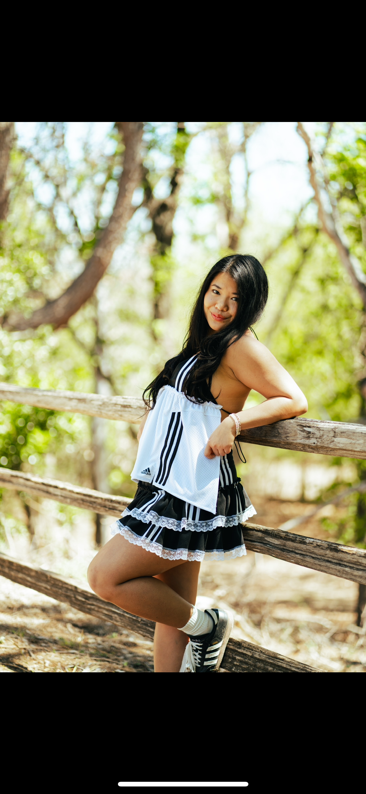 Young woman leaning on a wooden fence outdoors in a forested area, wearing black and white athletic clothing, sneakers, and holding a white mesh sports jersey.