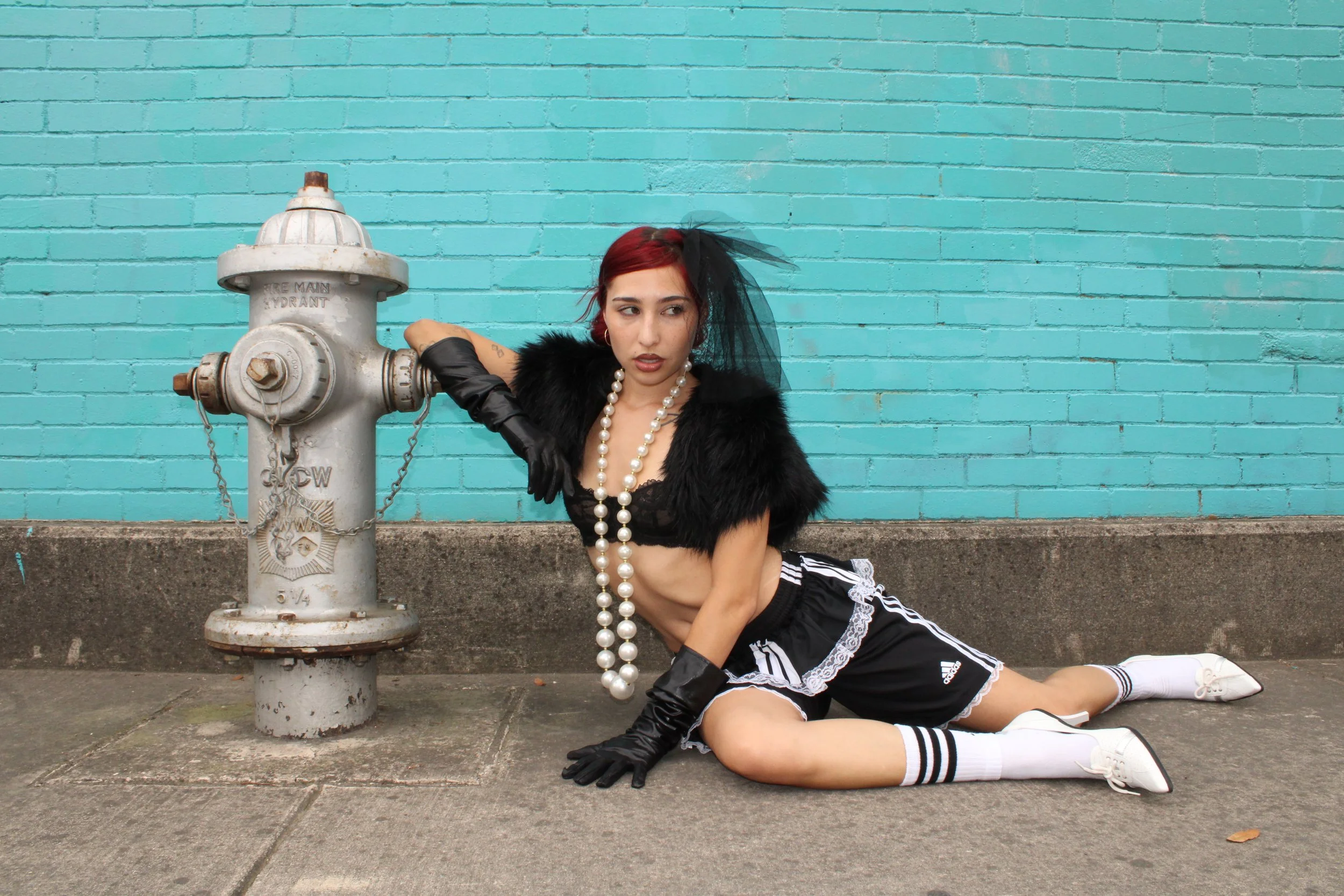 A young woman with red hair wearing a black and white athletic outfit, black gloves, and a pearl necklace, poses sitting on the sidewalk against a bright blue brick wall next to a fire hydrant. She is leaning on the hydrant with one arm, looking to h